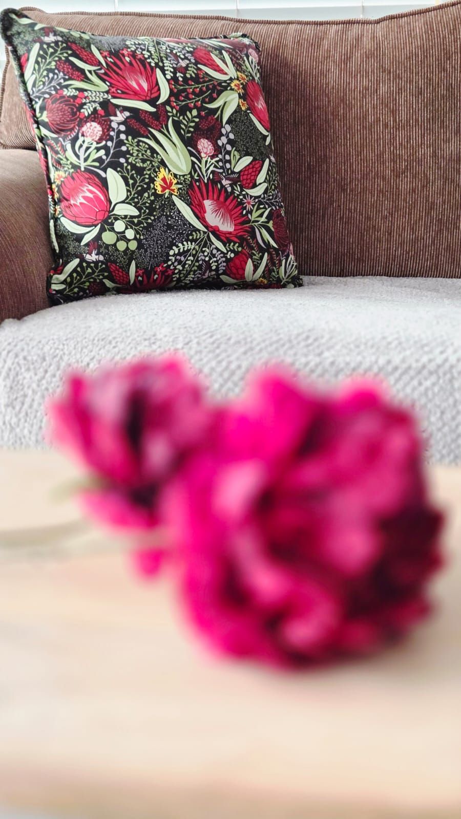 Pink peonies in soft focus on a wooden table with a floral cushion on a grey sofa in the background.