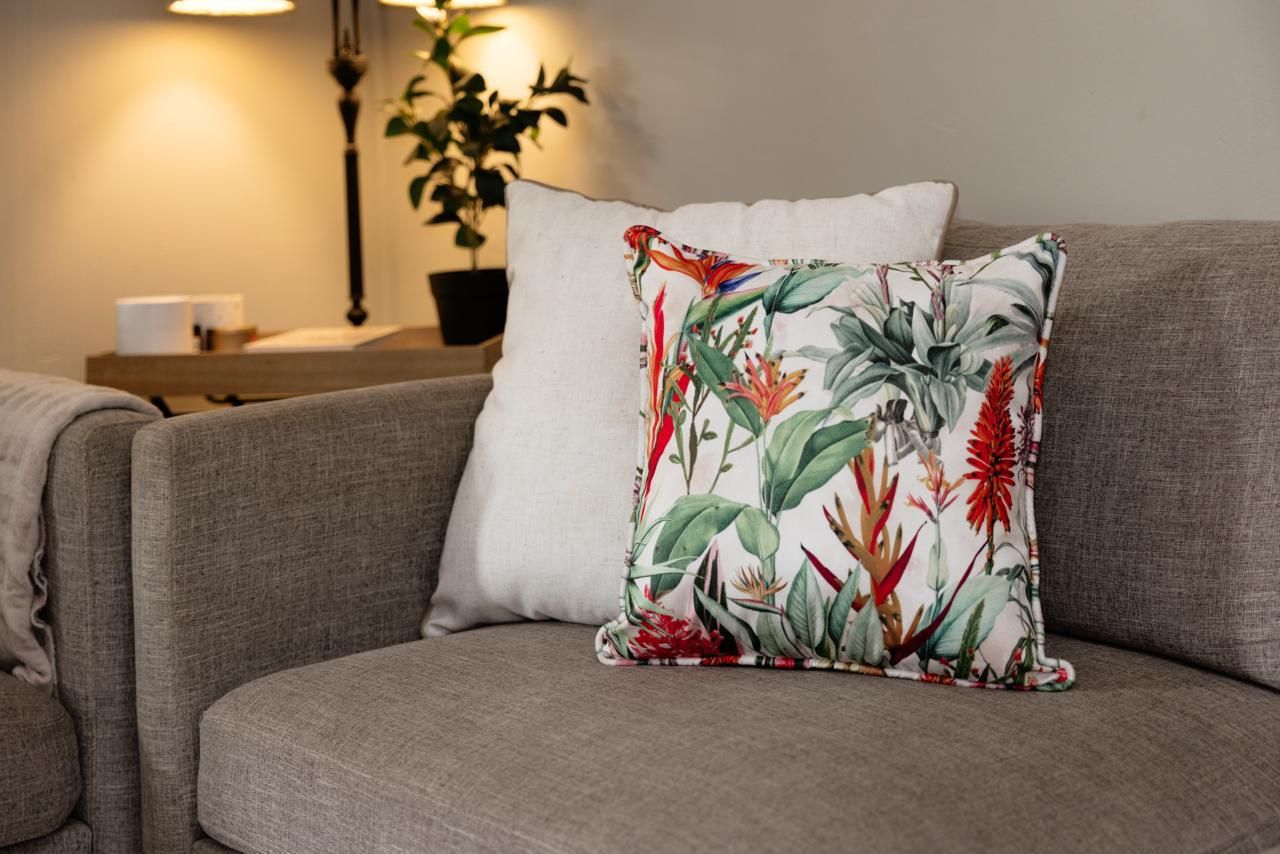 Decorative floral tropical print cushion on a grey sofa with white pillow and houseplant in background.