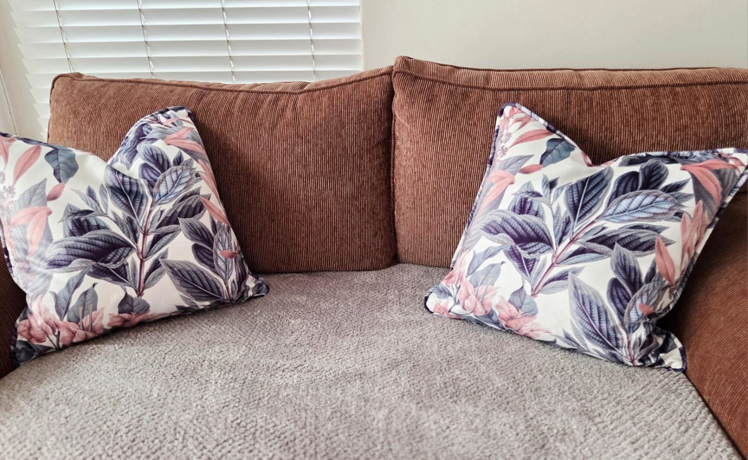 Brown sofa with two decorative floral throw pillows featuring purple leaves and pink accents in a bright living room.