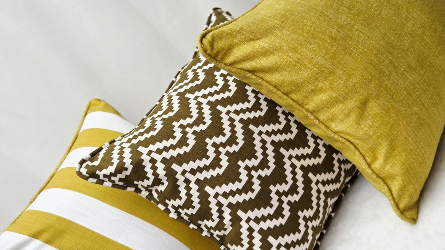 Three decorative pillows in gold and brown tones featuring striped, chevron, and solid patterns arranged on a white bed.
