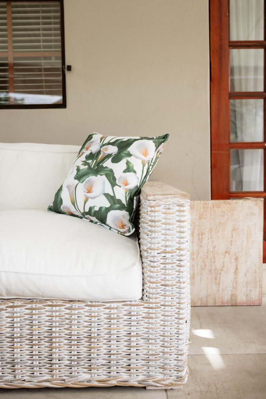 White wicker outdoor sofa with plush cushions and floral calla lily decorative pillow on a sunlit patio.