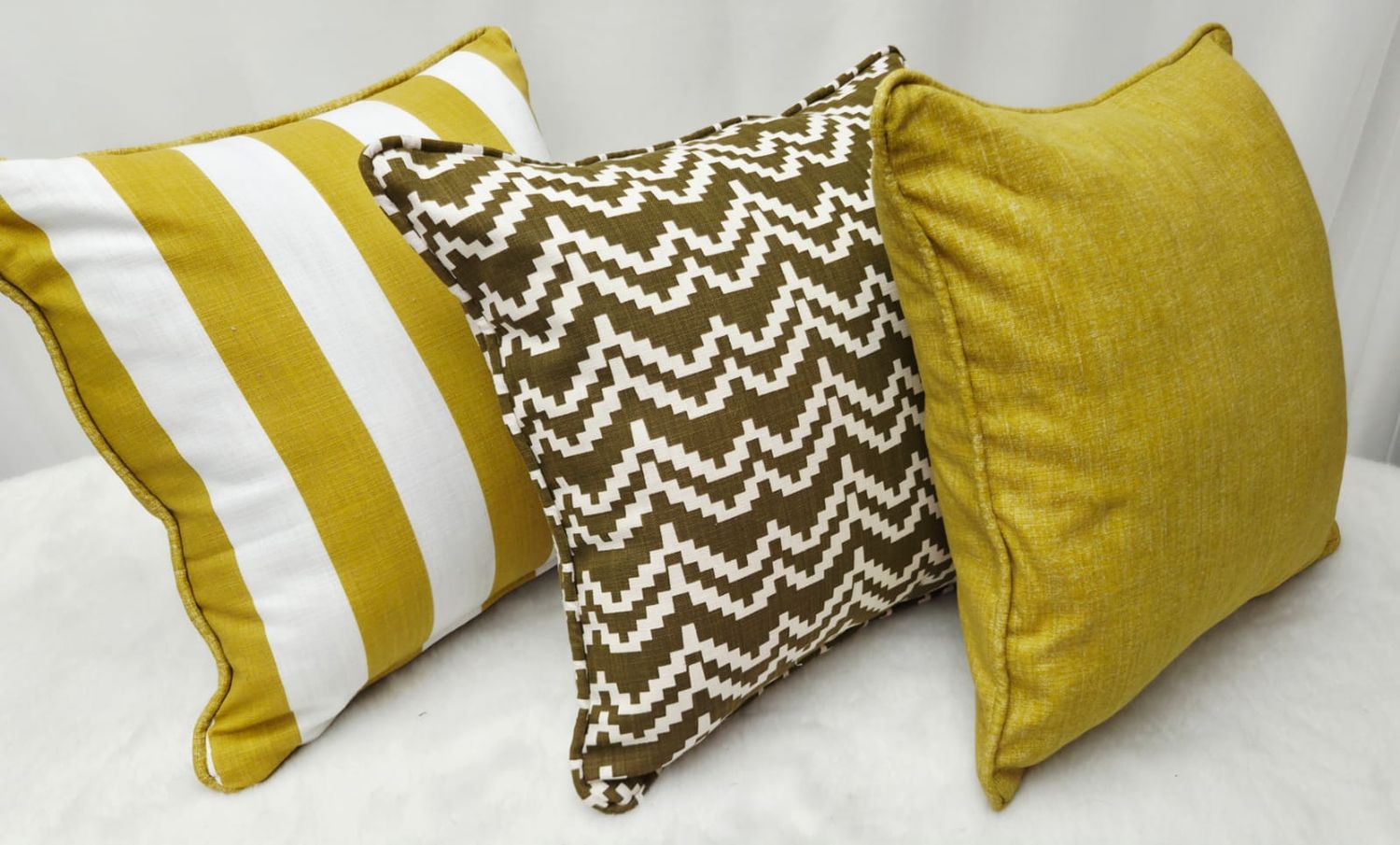 Three decorative throw pillows in gold and brown tones featuring stripe, chevron, and solid patterns on a white surface.