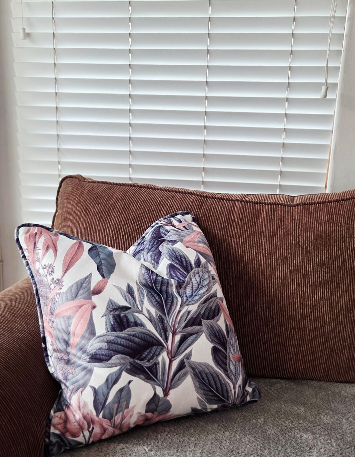 Decorative floral throw pillow with purple and pink leaves on a brown chenille sofa near white window blinds.