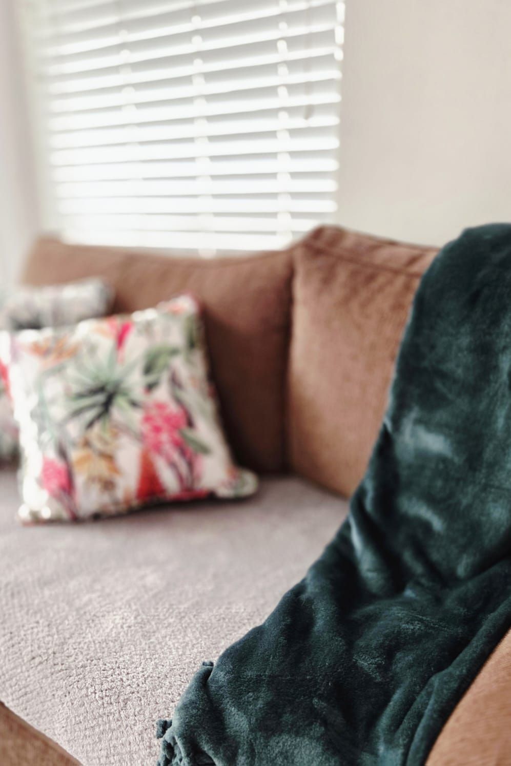 A cozy brown sofa with a floral throw pillow and green velvet blanket near a window with white blinds.