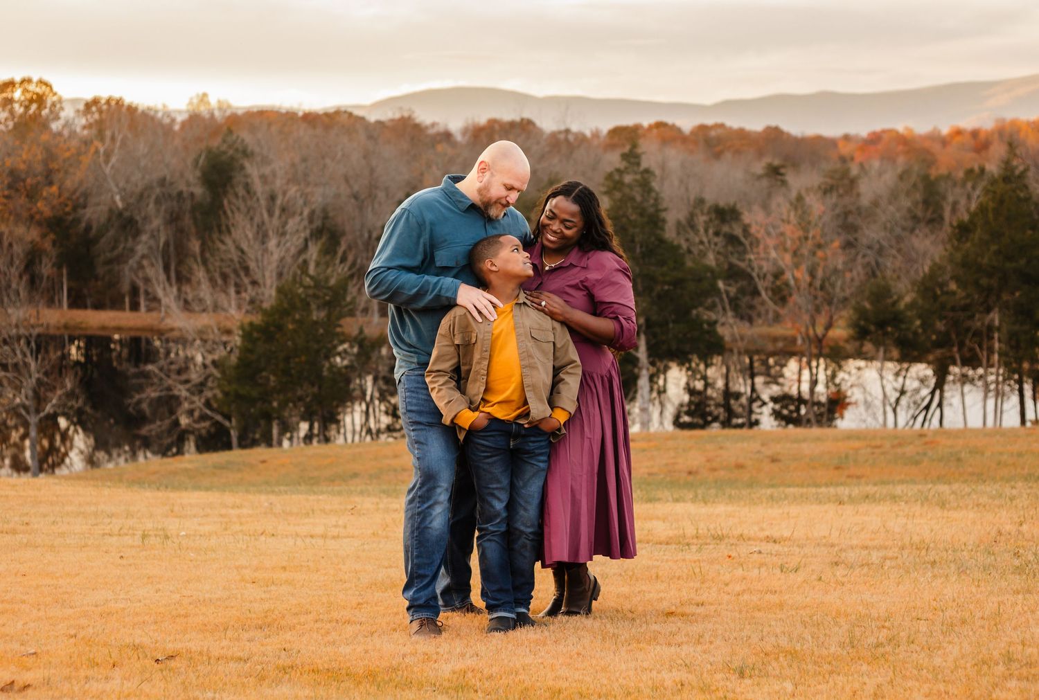 Family of three with cuddle together against a fall mountain backdrop for their Charlottesville photoshoot, with mom wearing purple dress.