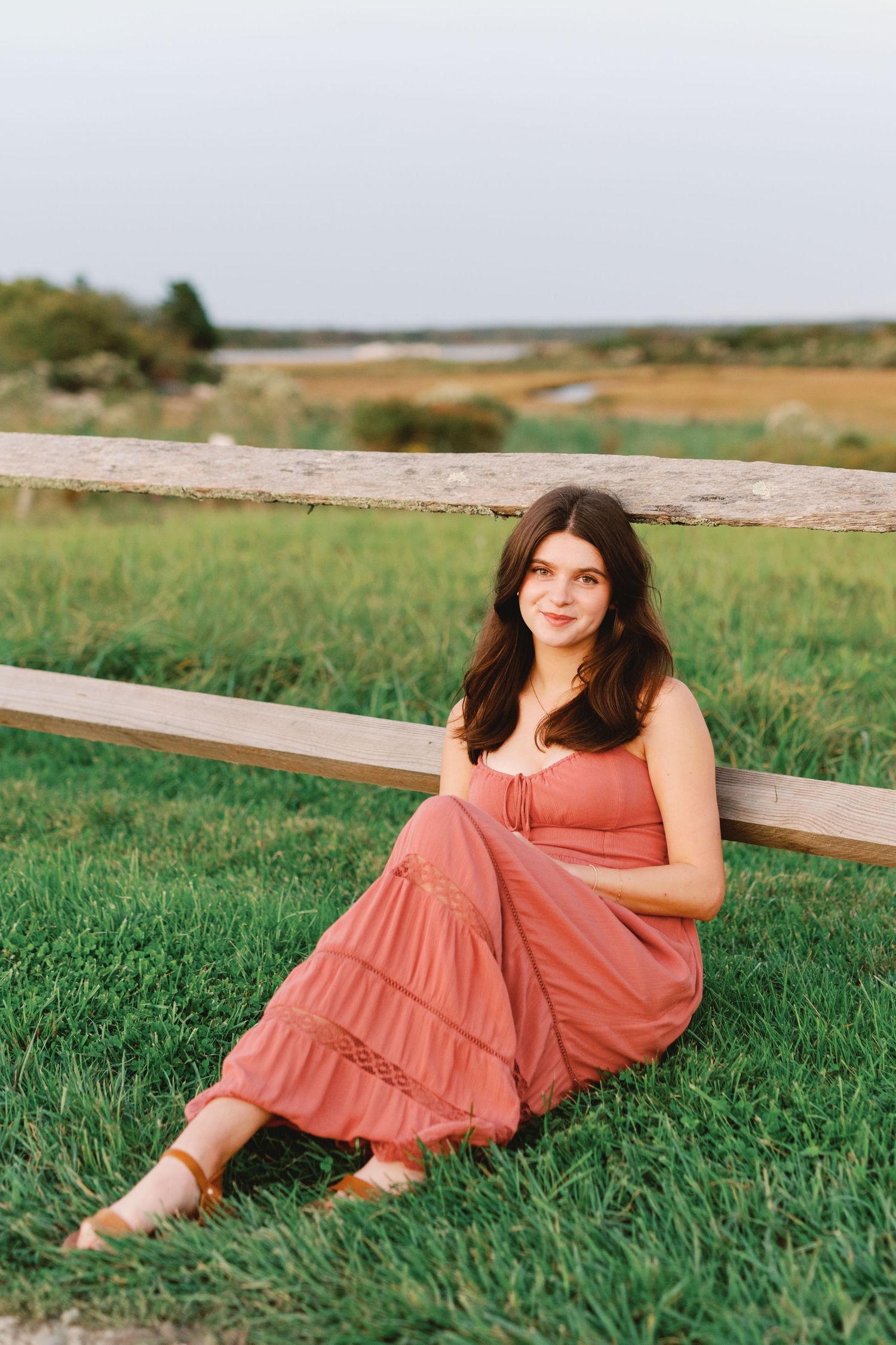 Young woman in pink maxi dress sitting on grass leaning against wooden fence in scenic outdoor field setting.