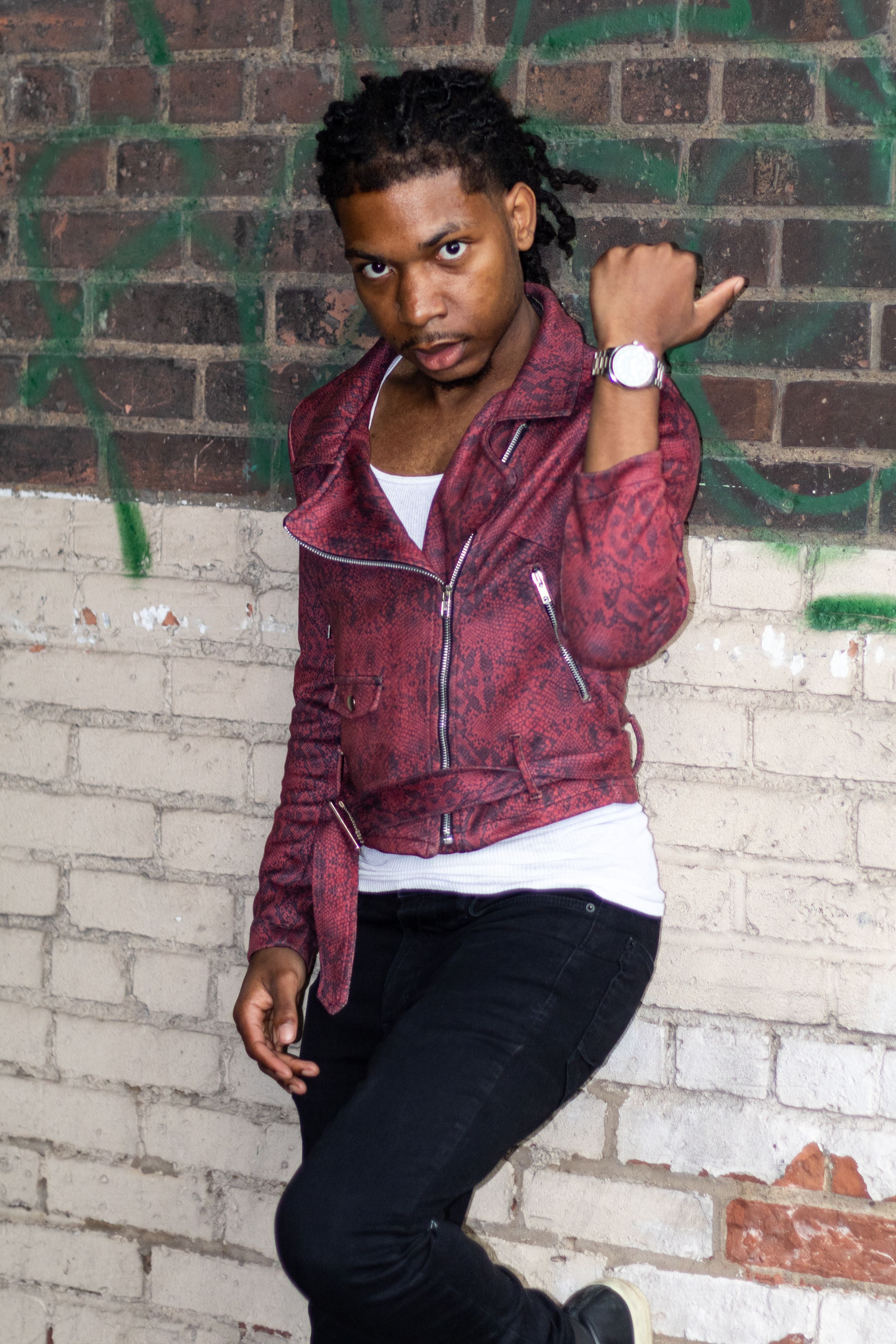 Young man in red jacket with dreadlocks posing against graffiti brick wall wearing a watch.
