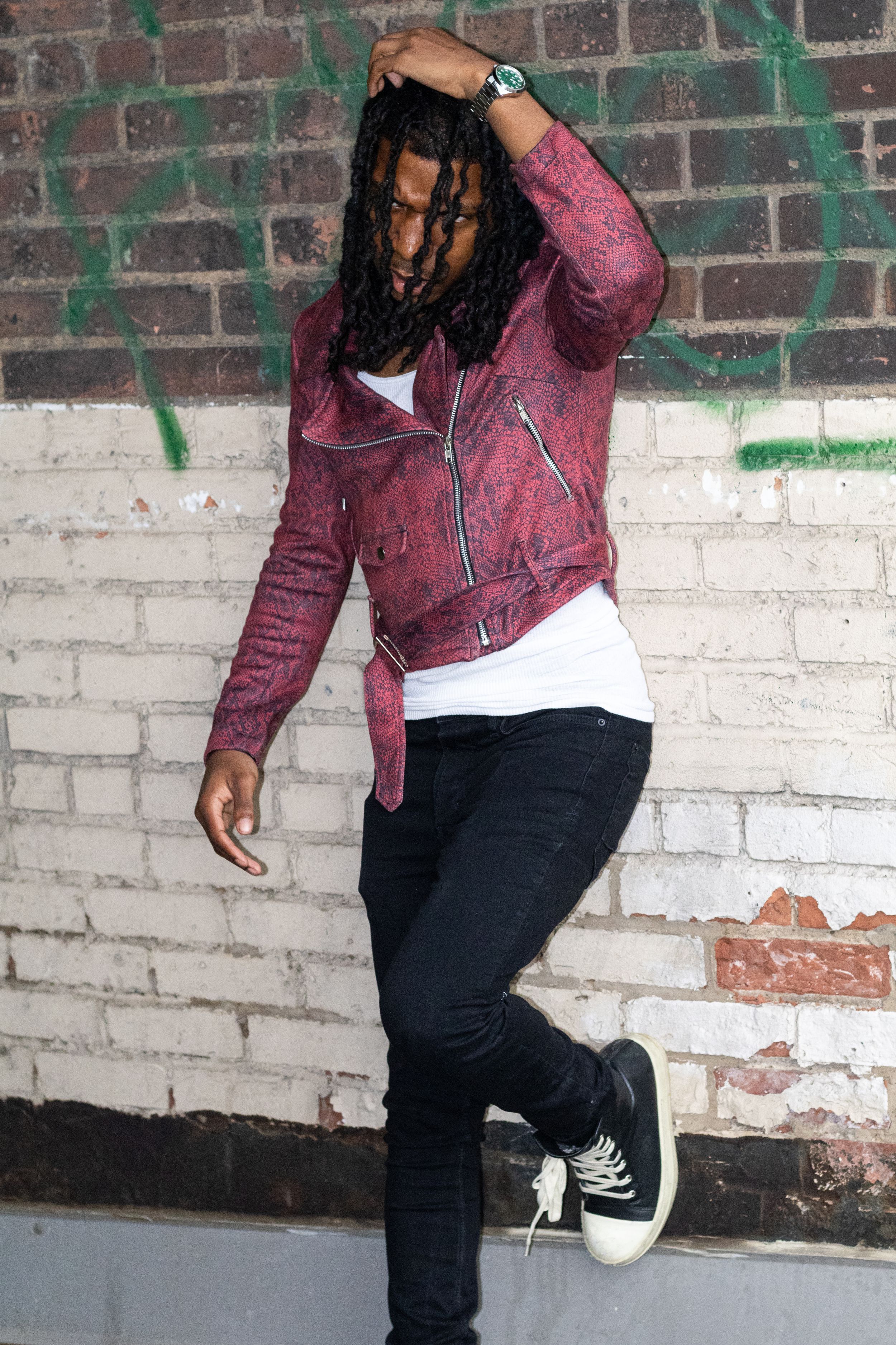 Young man with dreadlocks wearing red leather jacket and black jeans posing against a brick wall.