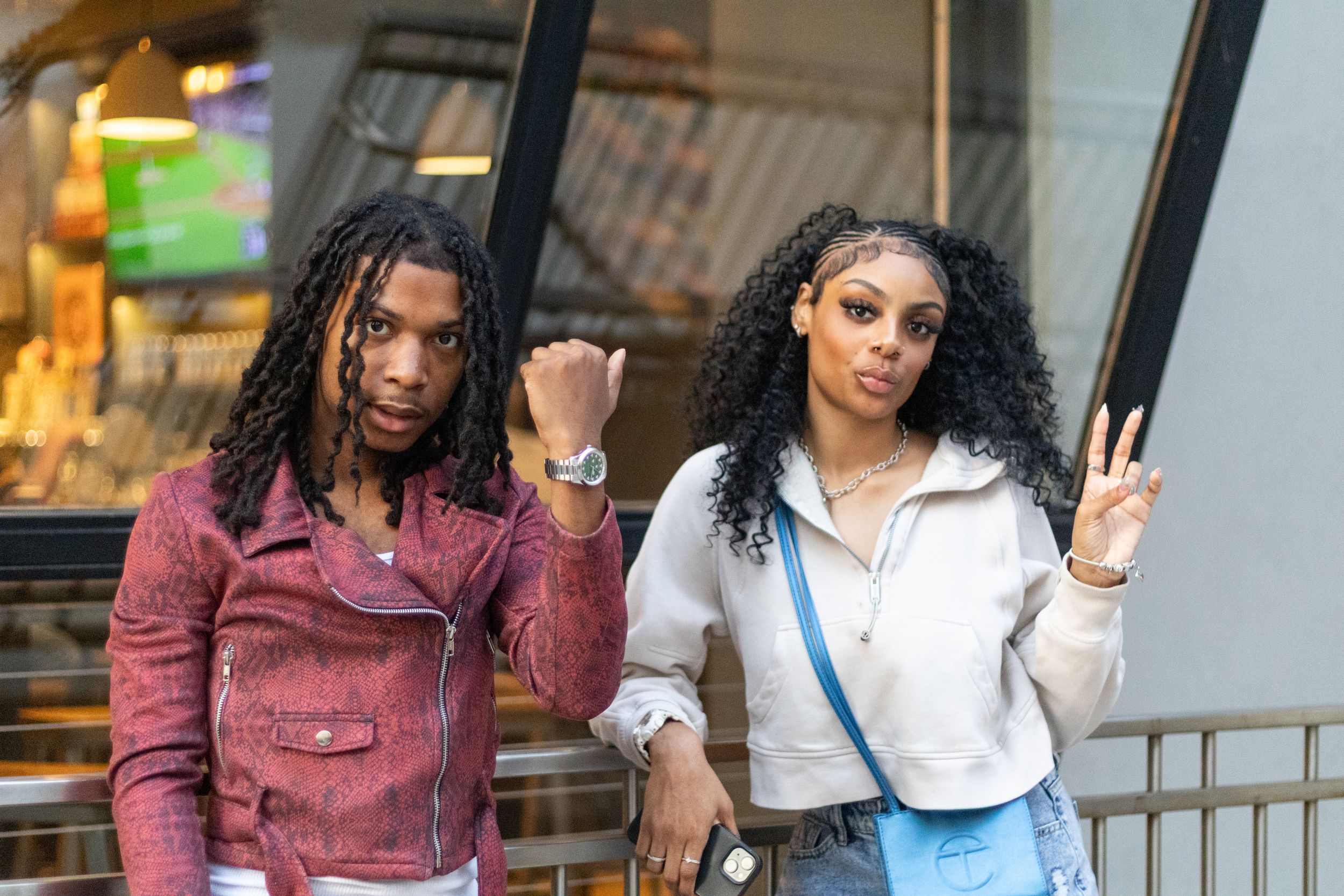Two young people posing outdoors, one showing a fist and the other a peace sign, wearing casual stylish outfits.