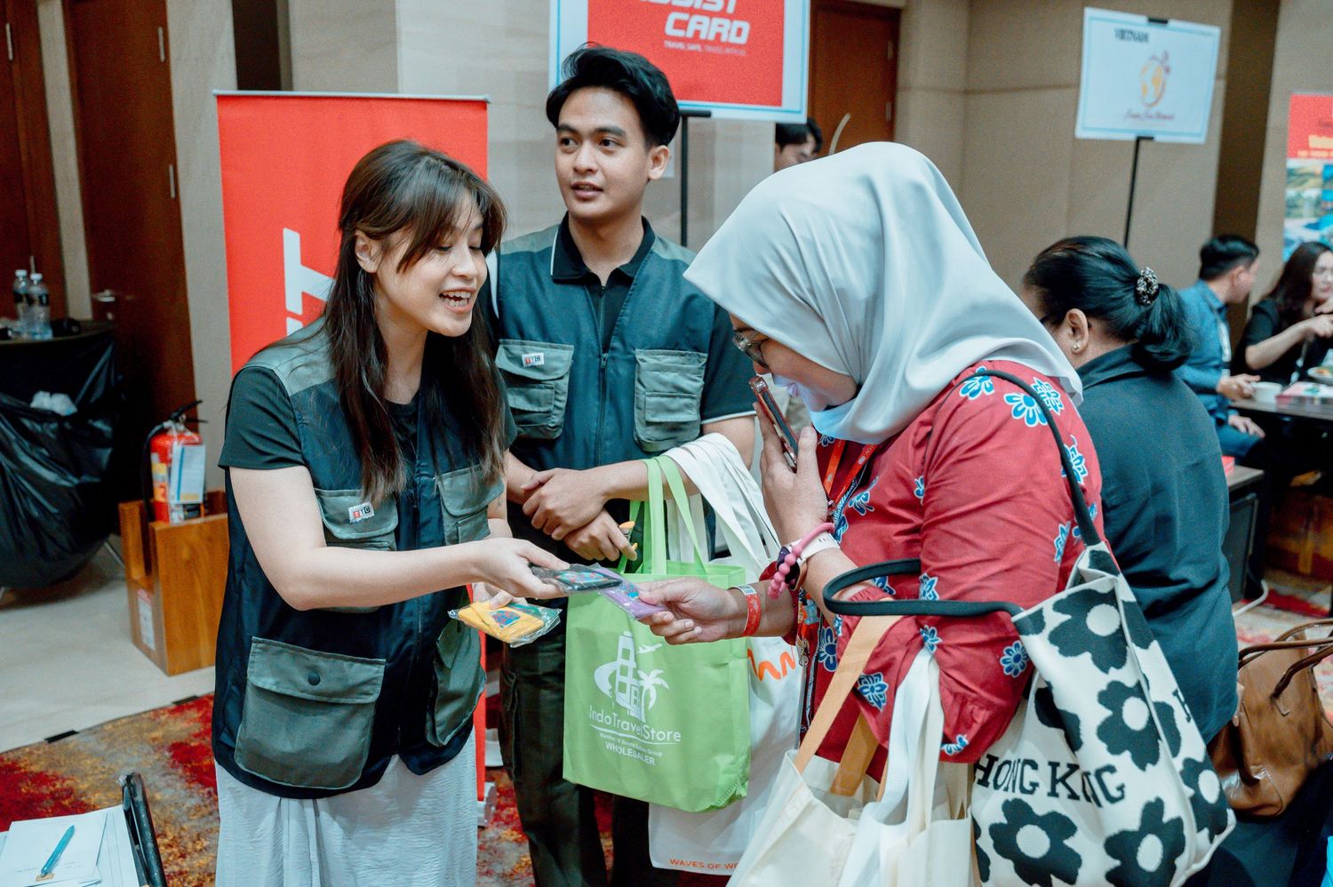 Two staff members in branded vests greet a visitor at an indoor promotional event or job fair.