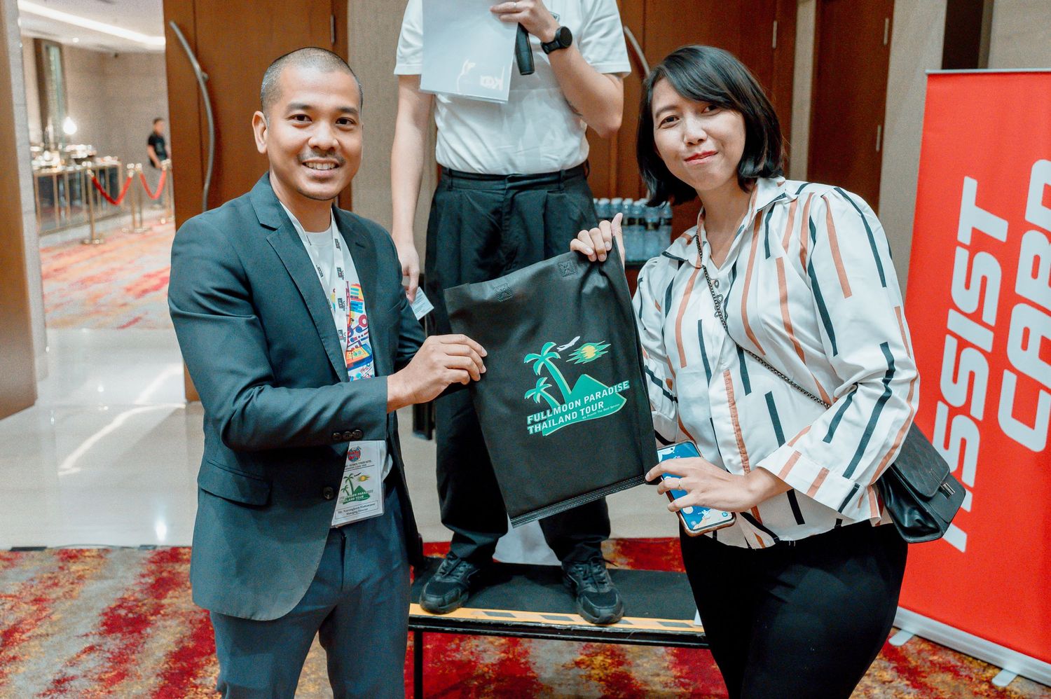 Two professionals exchange a branded tote bag at a corporate event, smiling in a modern hotel conference venue.