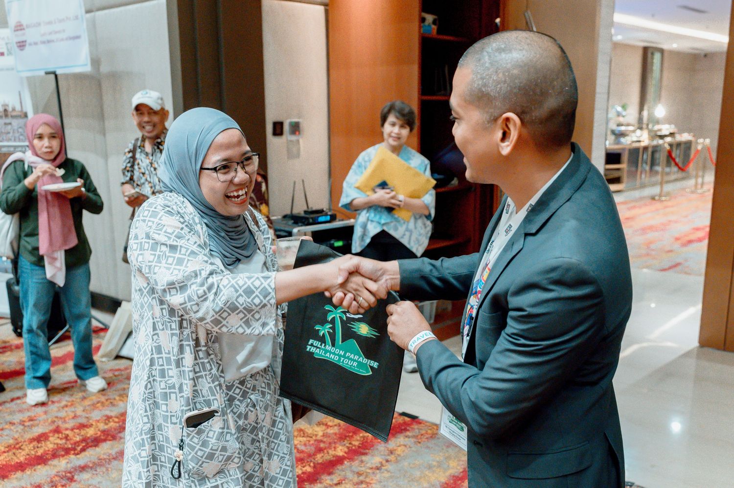 A woman in a hijab shakes hands with a man in a suit at a formal indoor event, exchanging a gift bag.