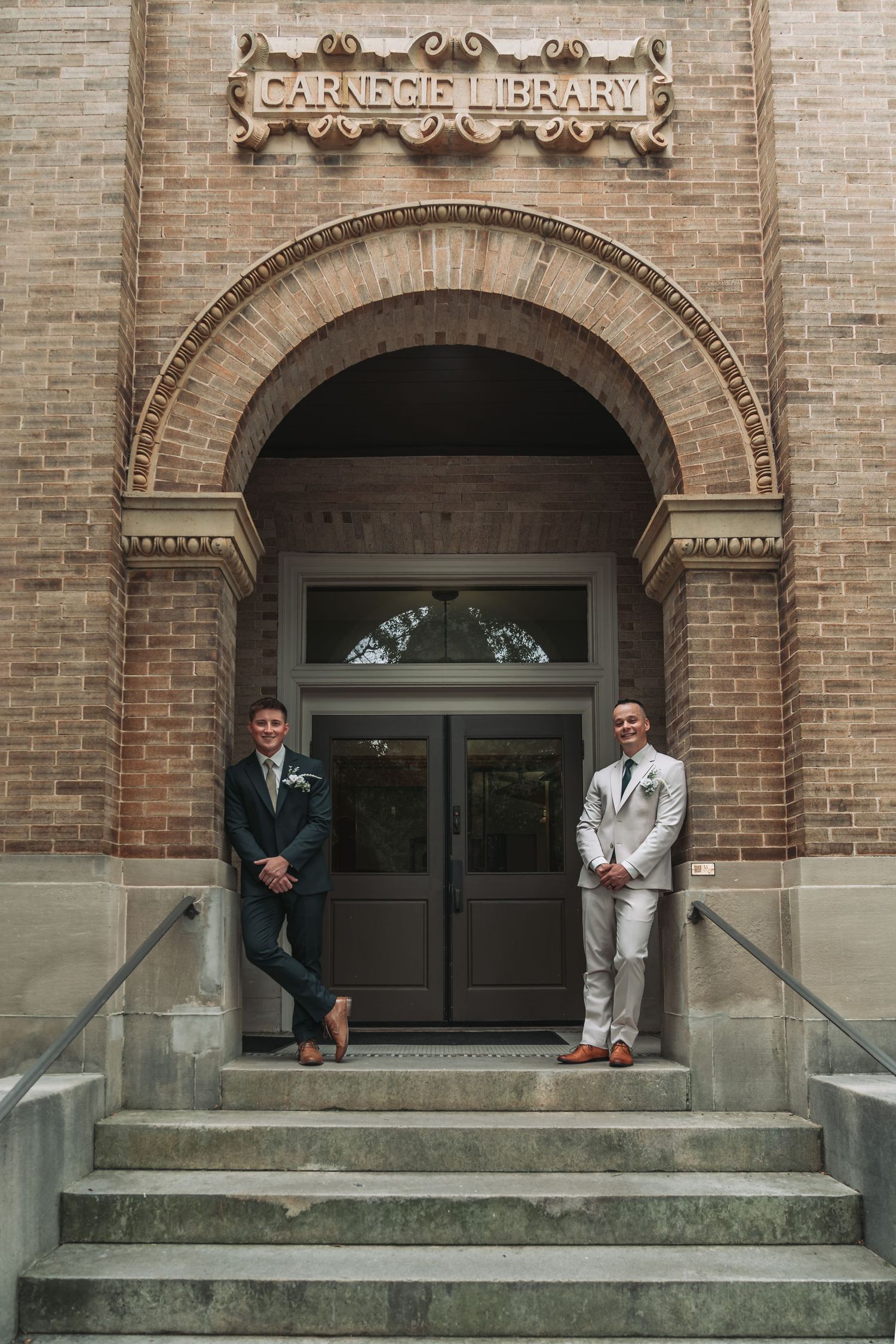 Black and white photo of two grooms standing on steps outside a brick building during their wedding ceremony.