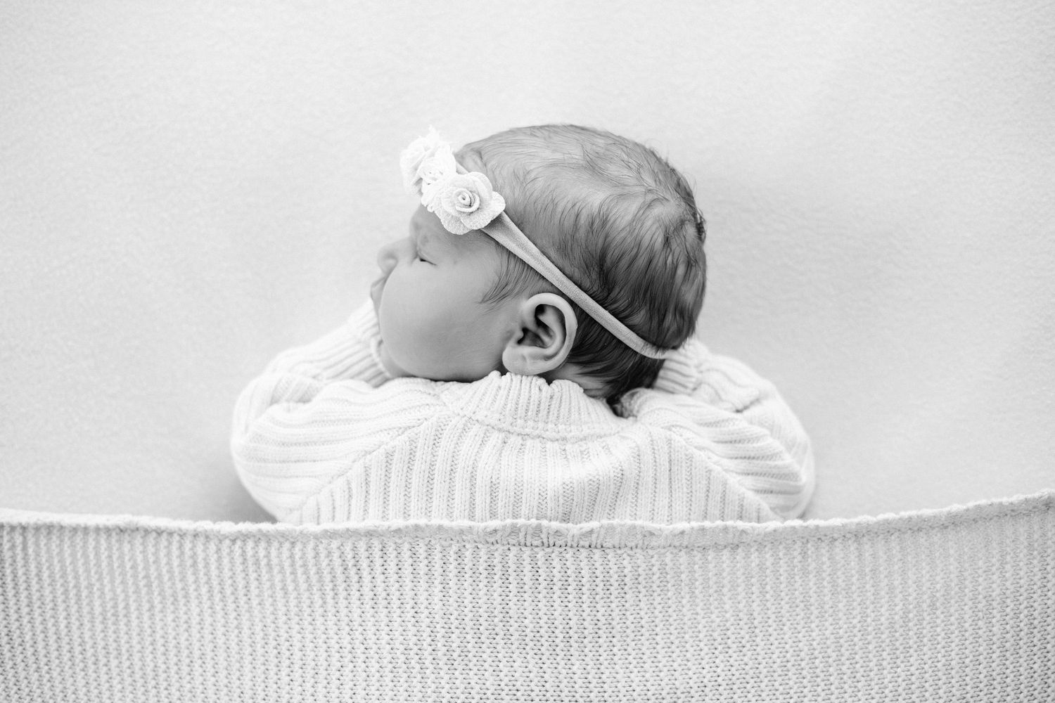 Black and white photo of a sleeping newborn baby wearing a floral headband, lying on a soft blanket.