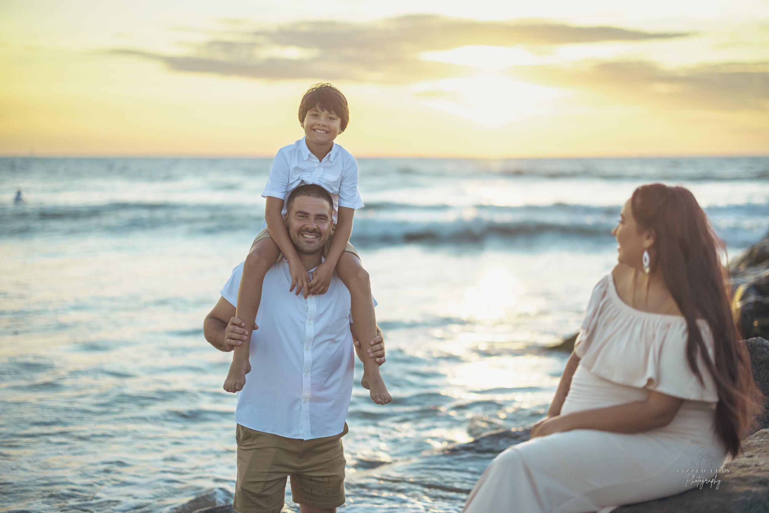 Family enjoying a beach sunset, father carrying son on shoulders, mother sitting on rocks, golden hour ocean backdrop.