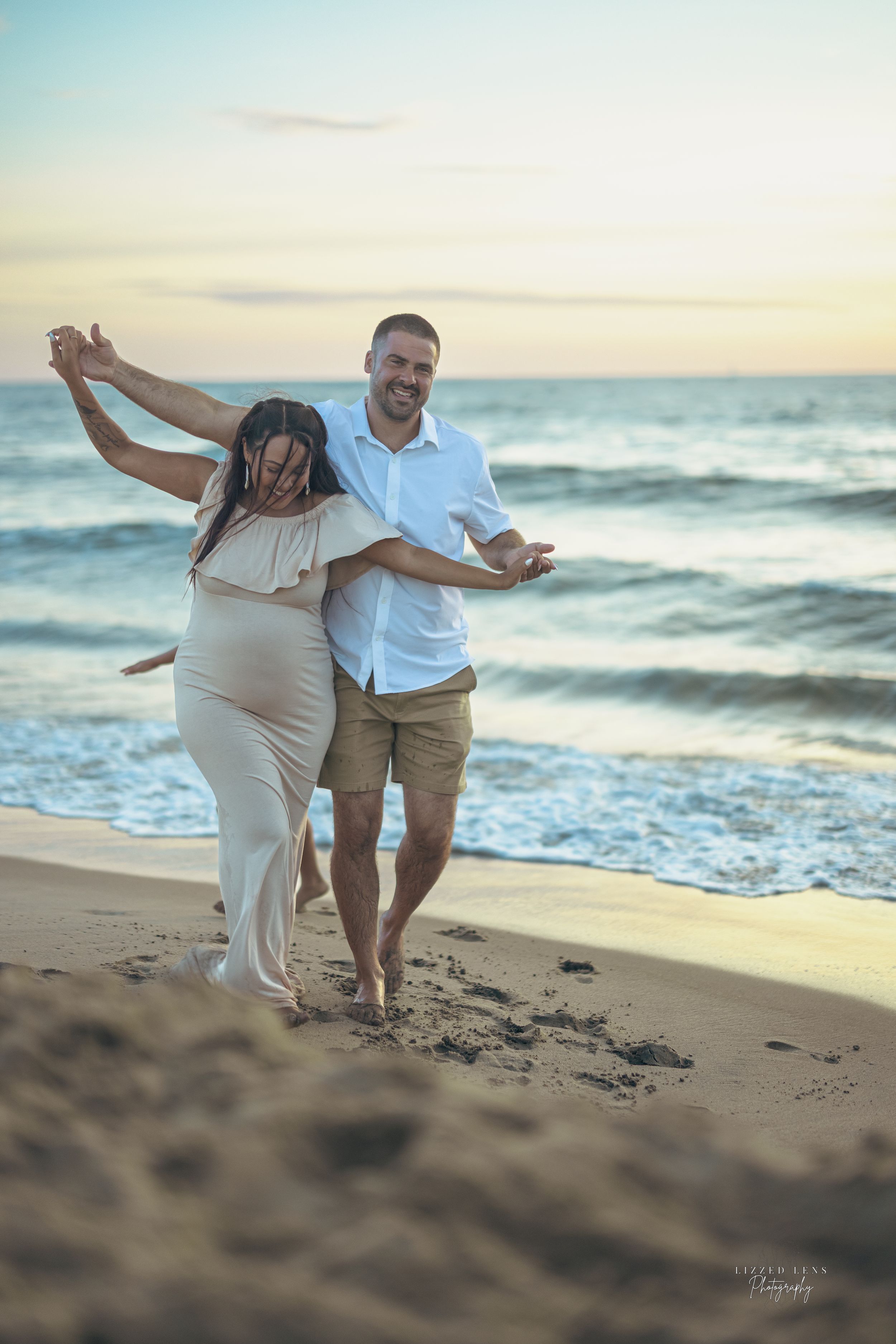 Happy couple dancing on sandy beach at sunset, pregnant woman in white dress with partner.