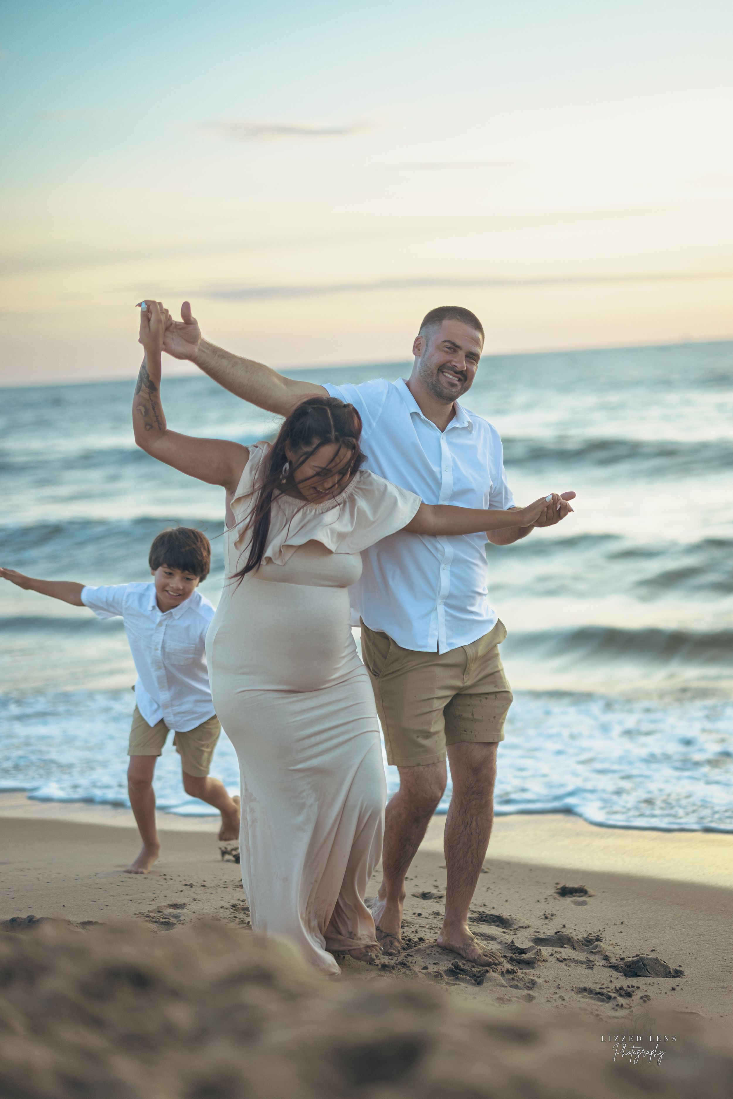 Pregnant woman dancing with partner and child on beach at sunset, family maternity photo session by the ocean.
