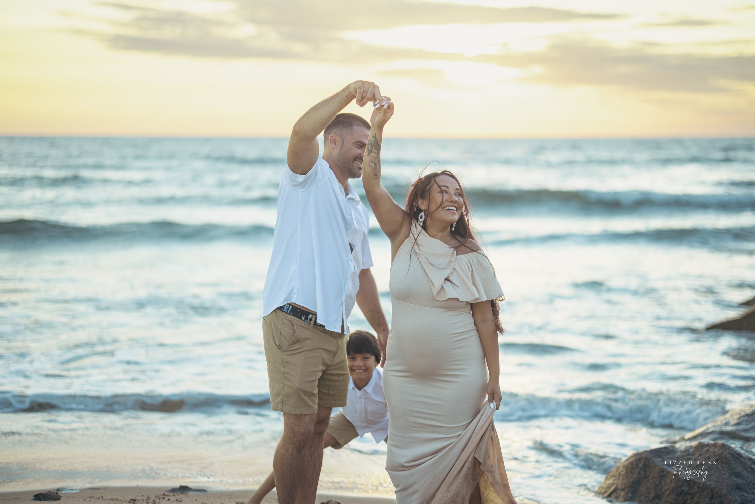 Pregnant woman in white dress dances with partner on beach at sunset, child visible in background.