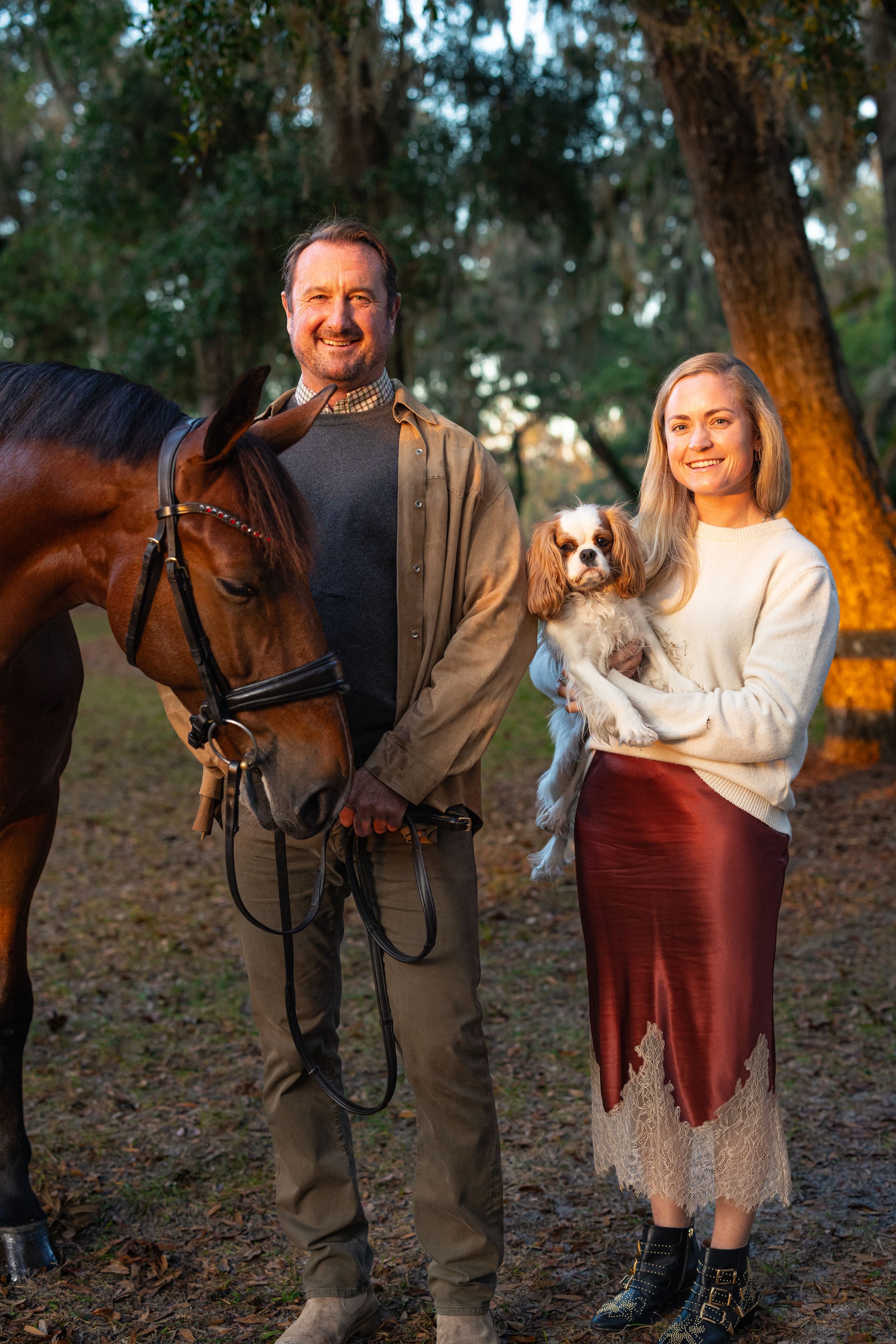 A smiling couple poses outdoors with a bay horse and a Cavalier King Charles Spaniel at golden hour.