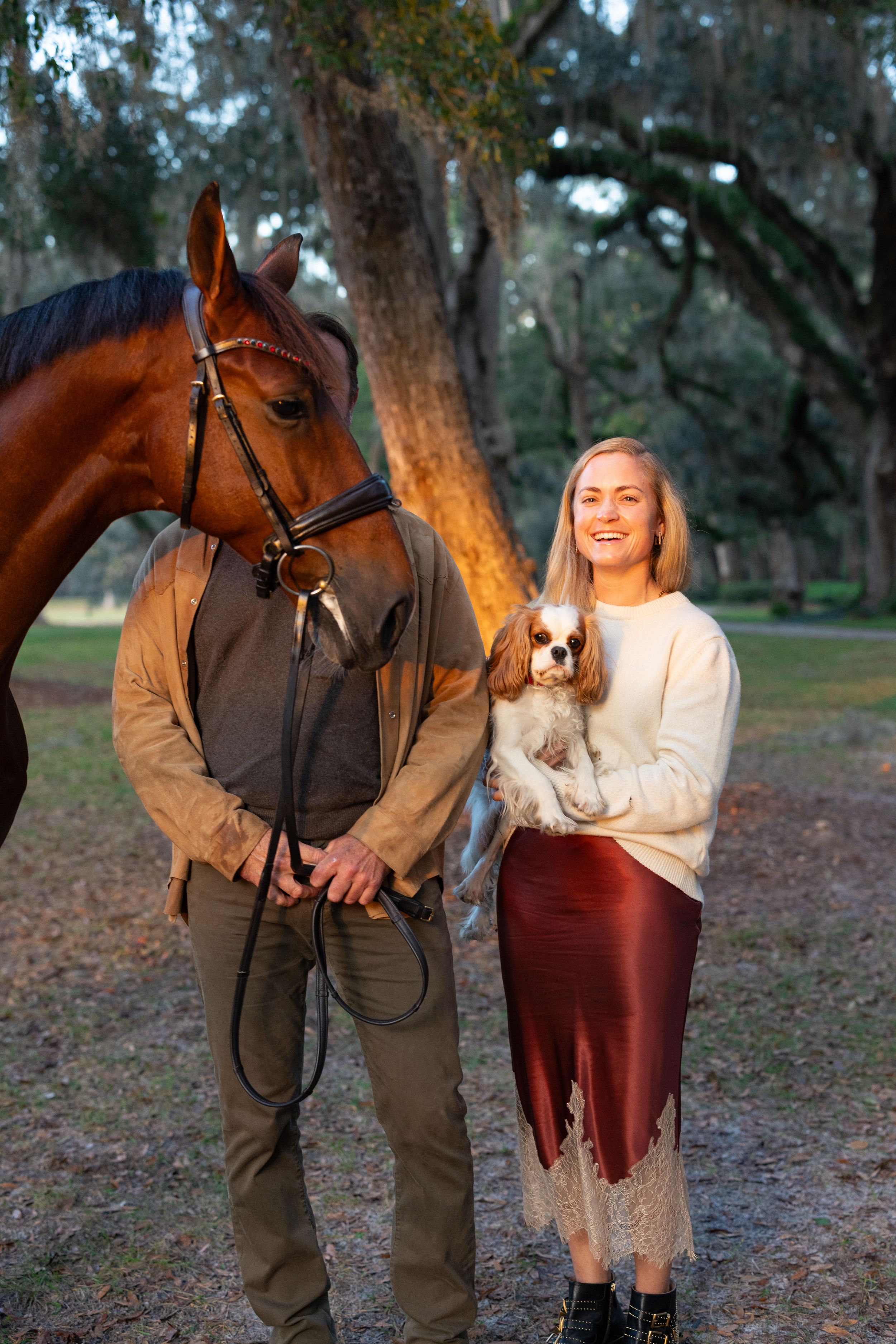 Smiling blonde woman holding a Cavalier King Charles Spaniel stands beside a bay horse and handler outdoors.