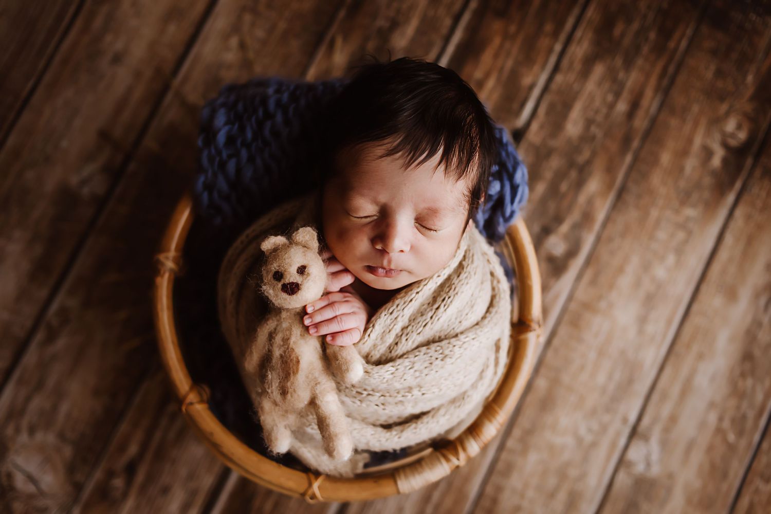 Newborn baby swaddled in cream wrap holding a teddy bear lovey, sleeping in a wooden basket on rustic wood floor.