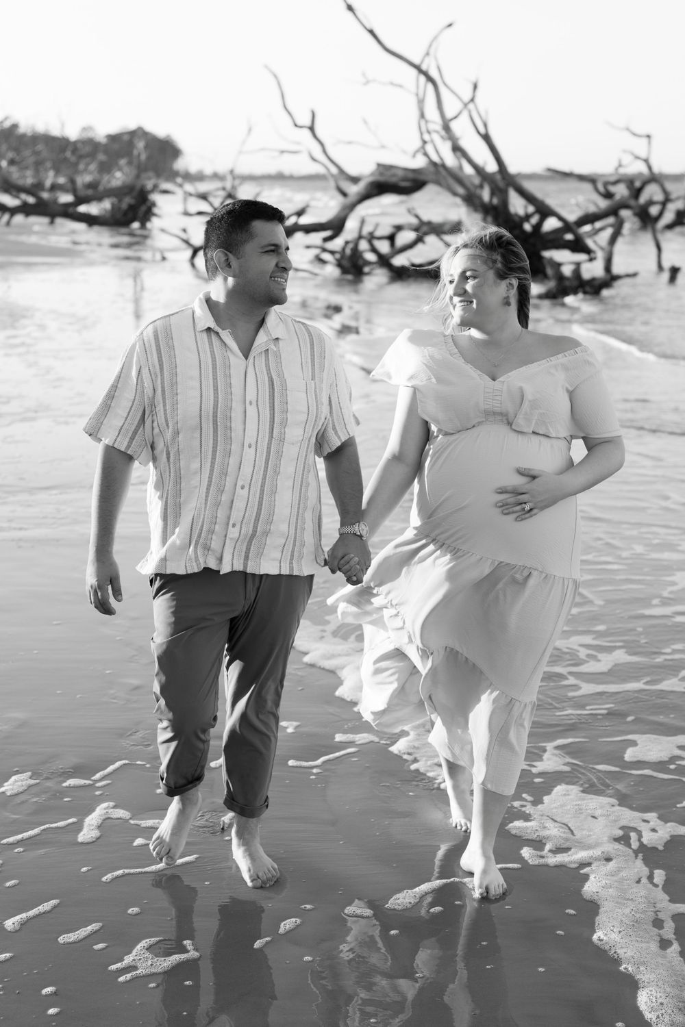Expectant parents walking hand in hand along Driftwood Beach in a black-and-white maternity portrait.
