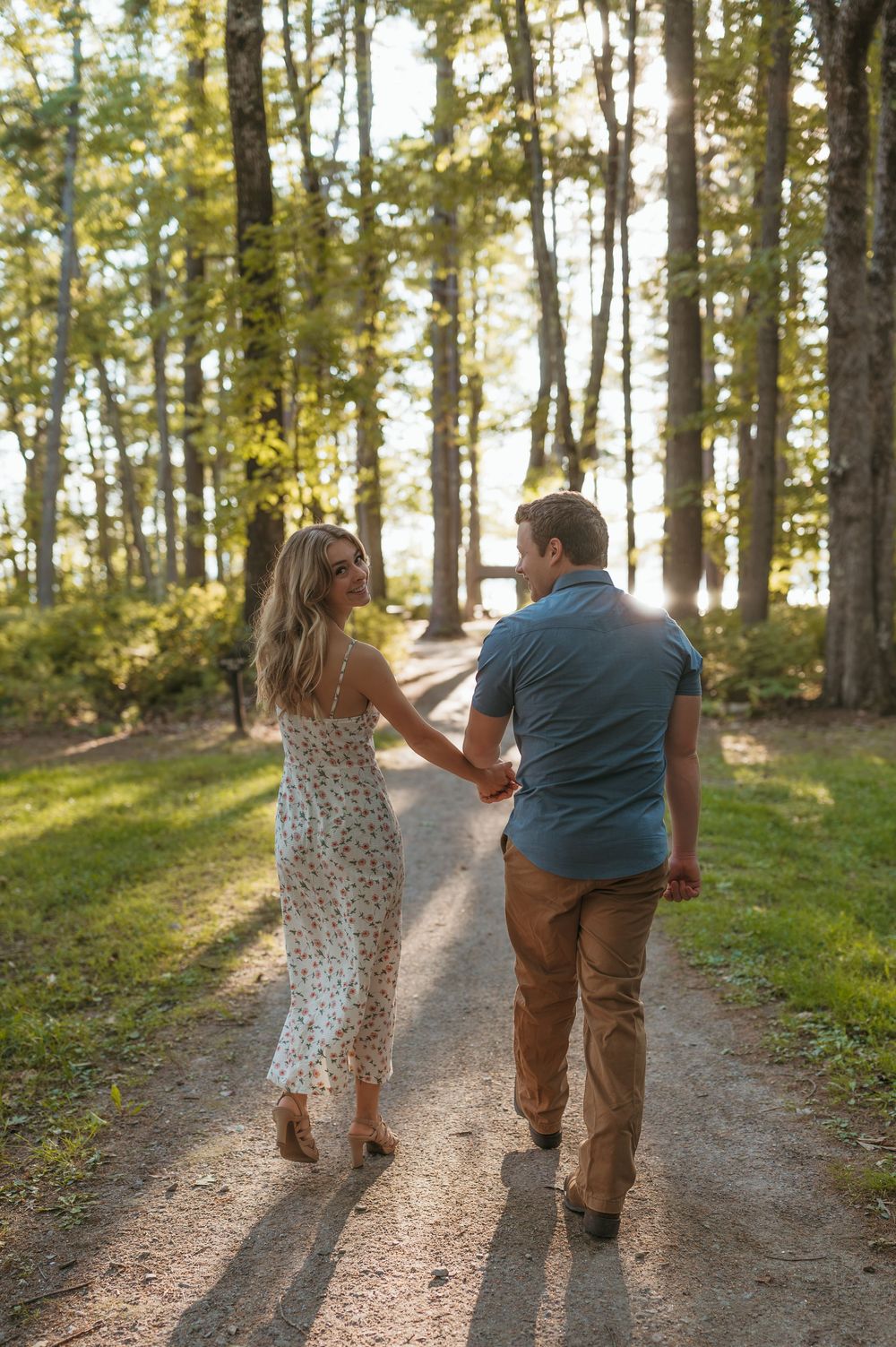 Couple holding hands walking down a sunlit forest path, woman in floral dress, man in blue shirt.