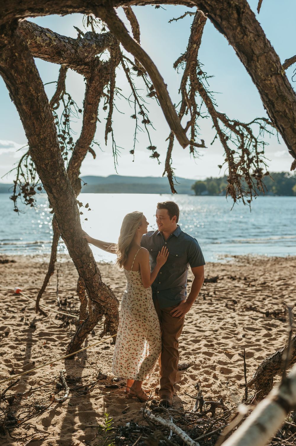 Couple sharing a romantic moment under a rustic tree on a sandy beach with calm blue water in the background.
