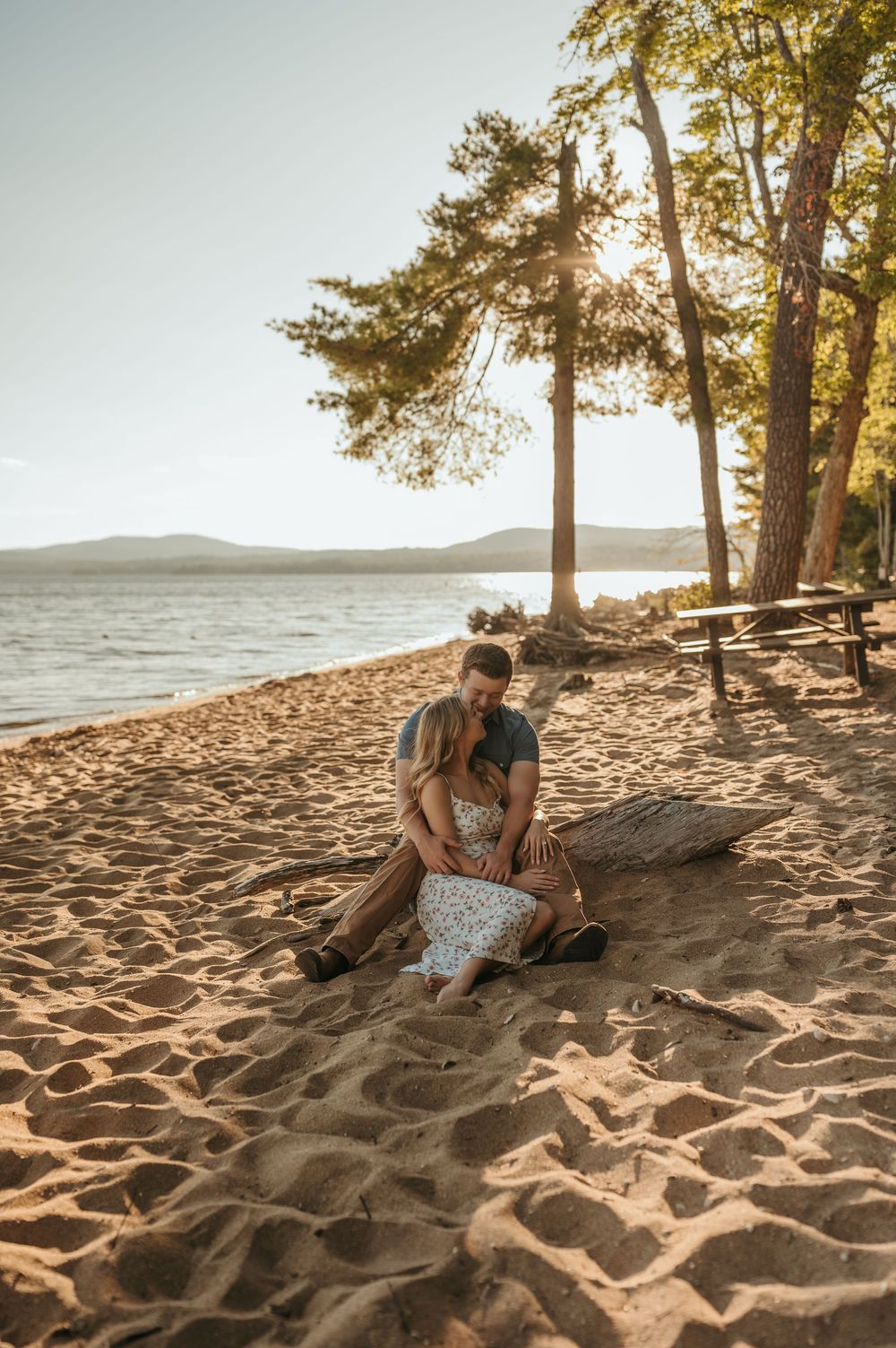 Couple sitting close together on a sandy beach at golden hour with trees and a lake in the background.