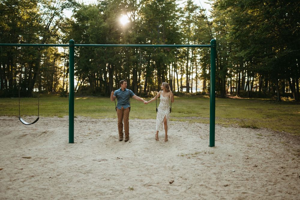 Couple holds hands on sandy playground with swing set, surrounded by tall trees in golden sunset light.