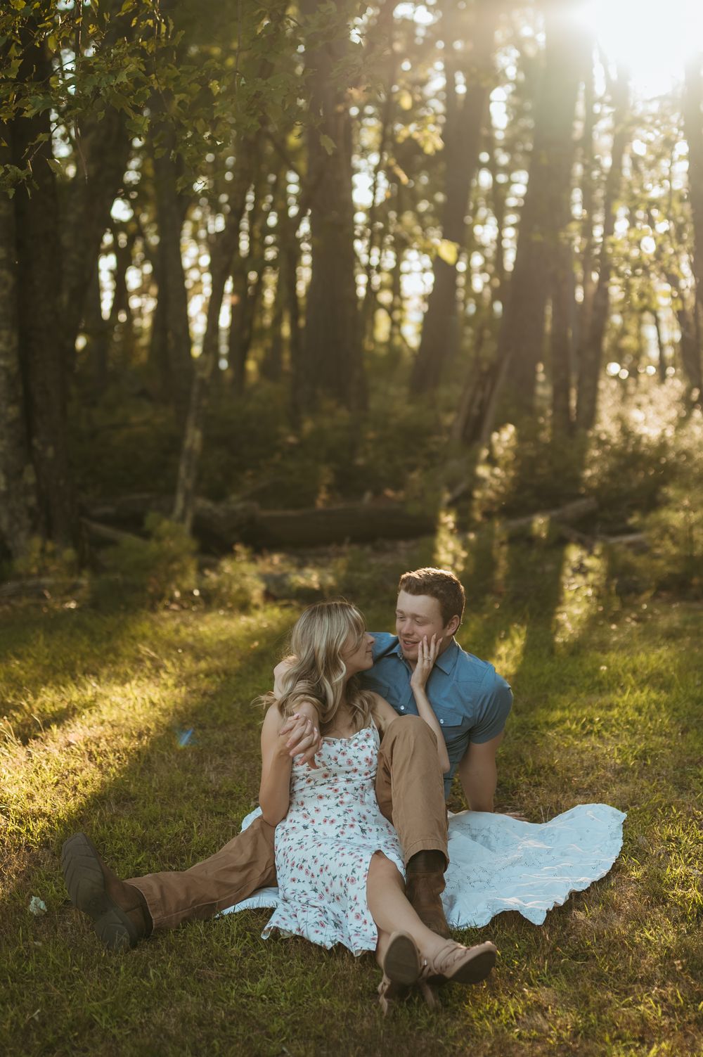 Couple sitting on grass in sunlit forest, woman in floral dress and boots, man in blue shirt, sharing a romantic moment.