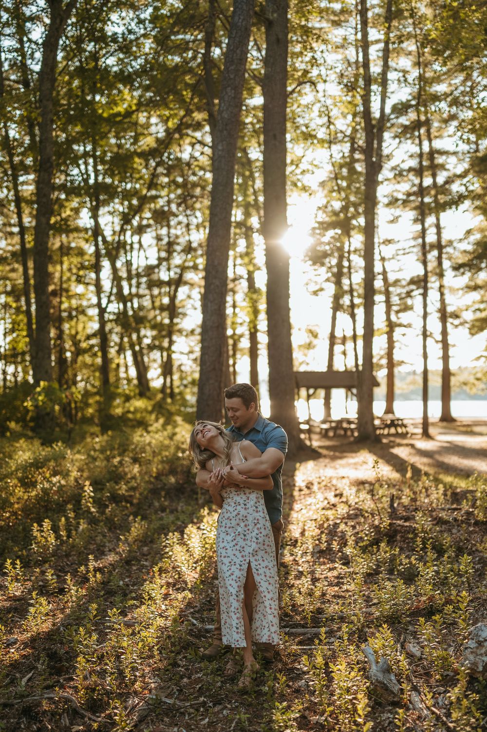 Couple embracing in sunlit forest near lake, golden hour light filtering through tall trees, woman in floral dress.