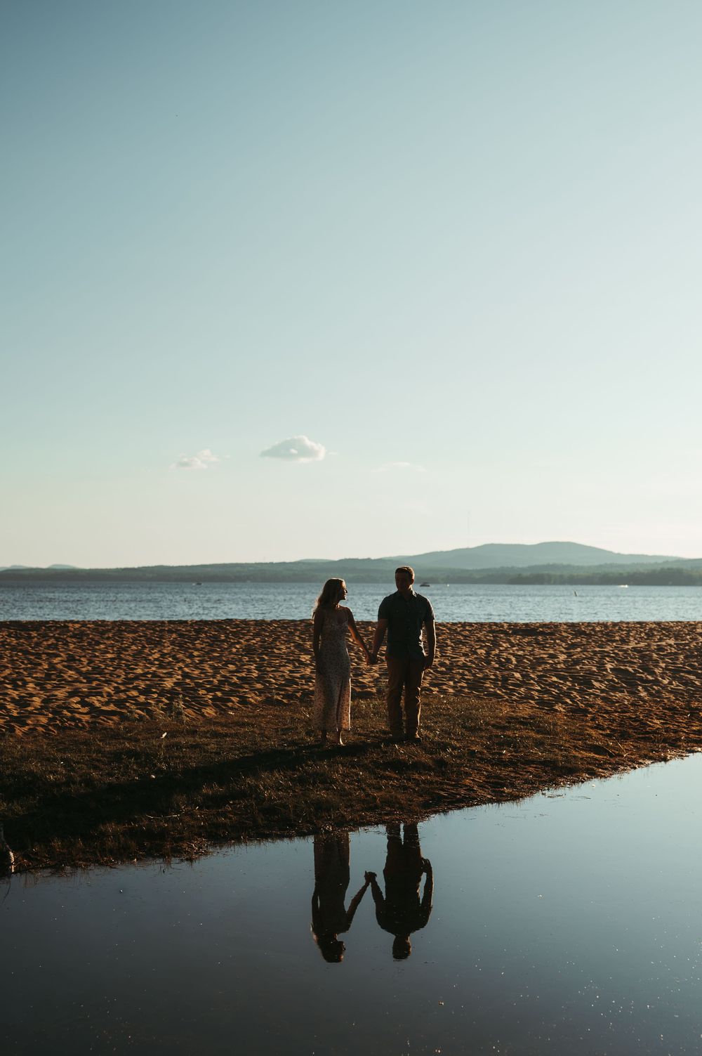 Couple holding hands on a sandy beach at sunset, with their reflection visible in a tidal pool, mountains in the background.