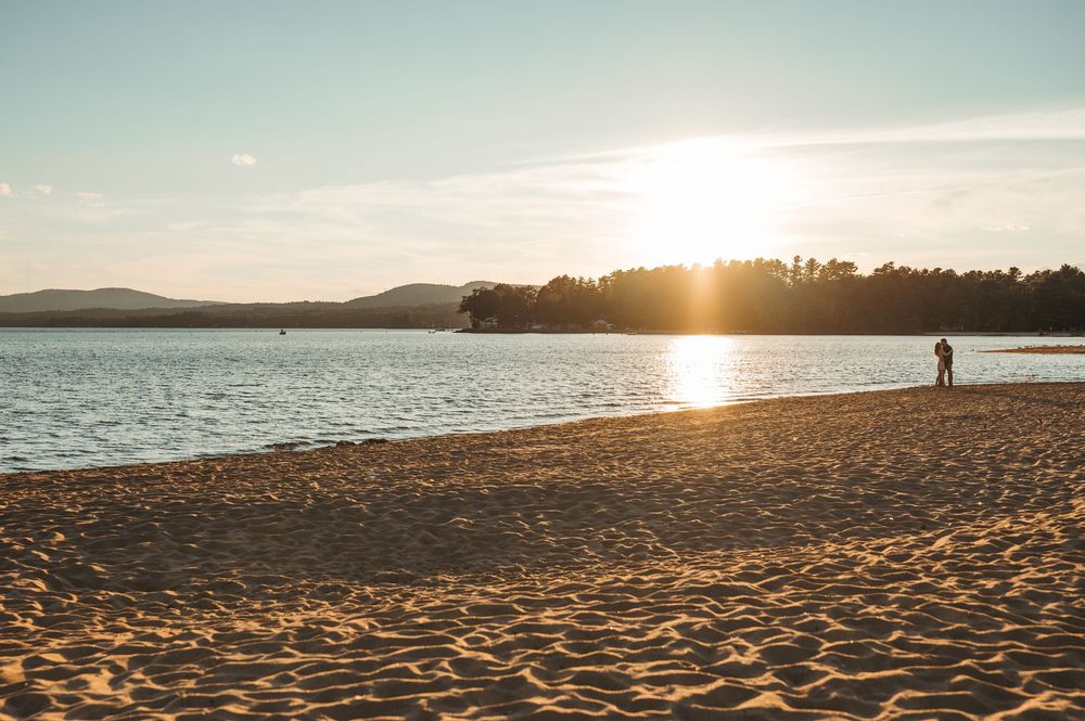 Golden sunset over a sandy beach with calm water, silhouetted trees, and a lone figure standing near the shoreline.