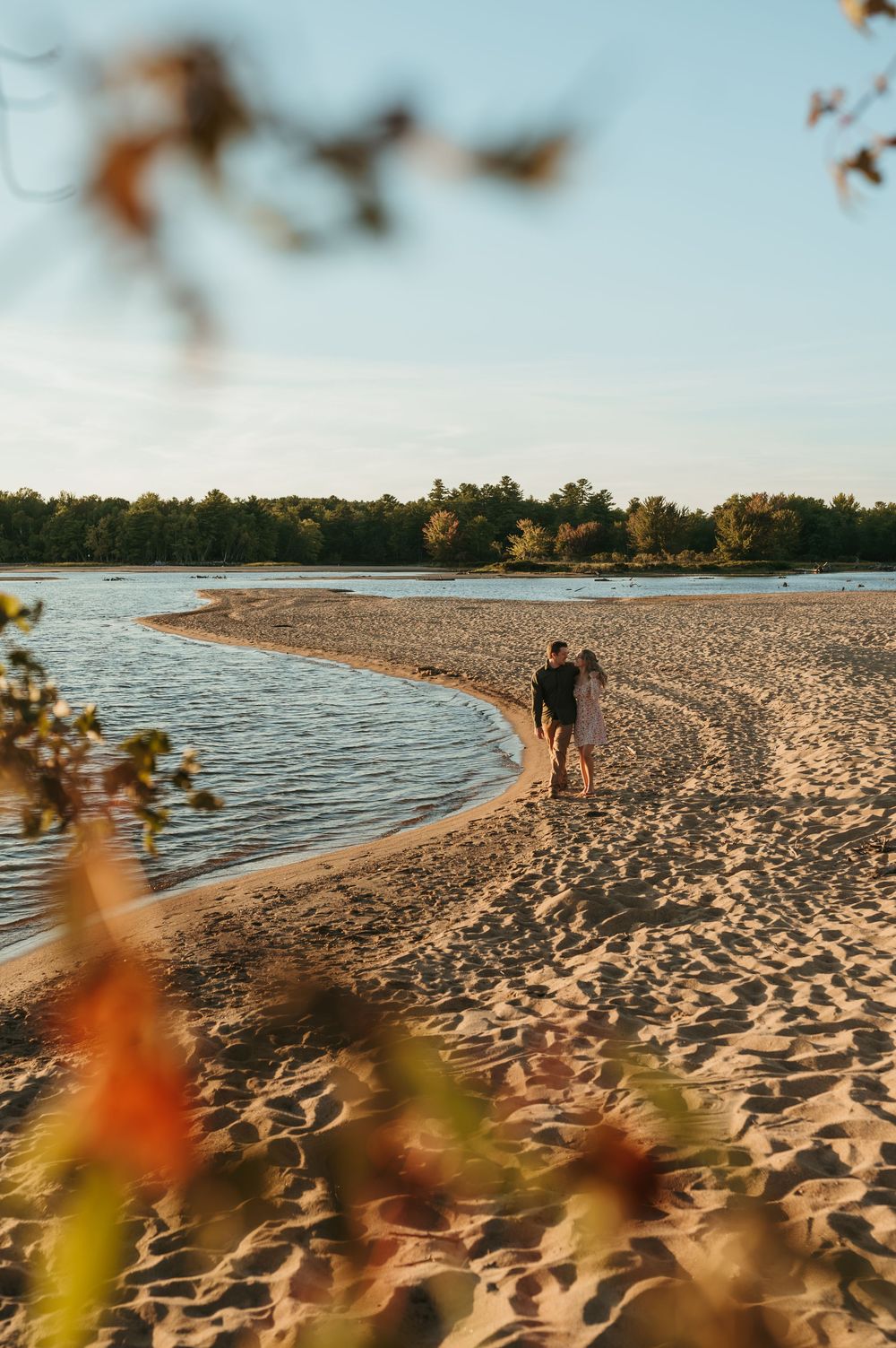 Couple walking hand in hand along a sandy beach shoreline at golden hour with trees in the background.