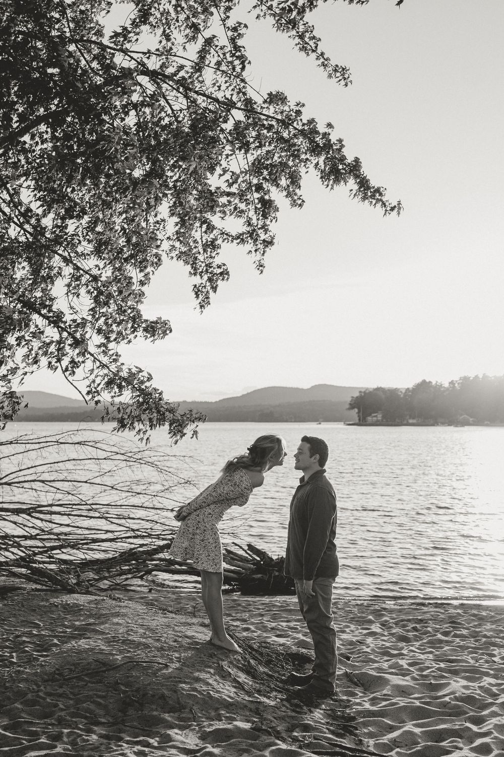 Black and white photo of a couple about to kiss on a sandy lakeside beach beneath overhanging tree branches.