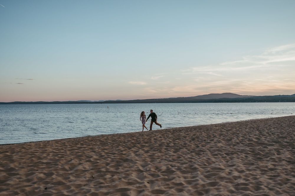 Two people hold hands and walk along a sandy beach at sunset with calm water and distant hills in the background.
