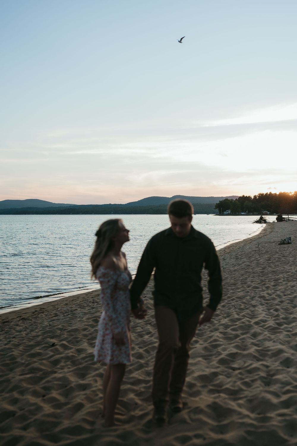 Couple holding hands walking on sandy beach at sunset with mountains and calm water in the background.