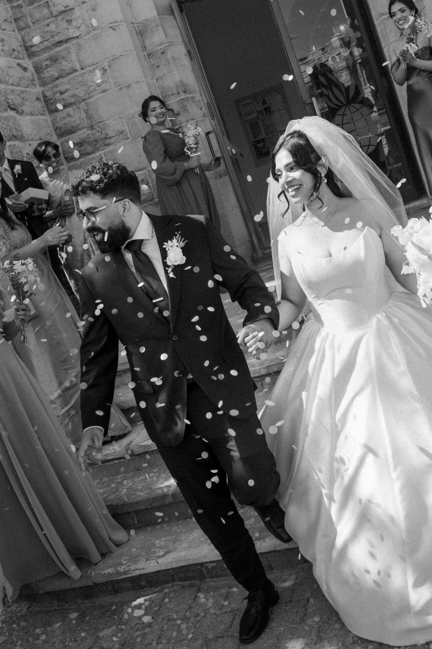 Black and white photo of a joyful bride and groom exiting church amid confetti shower, guests celebrating in background.