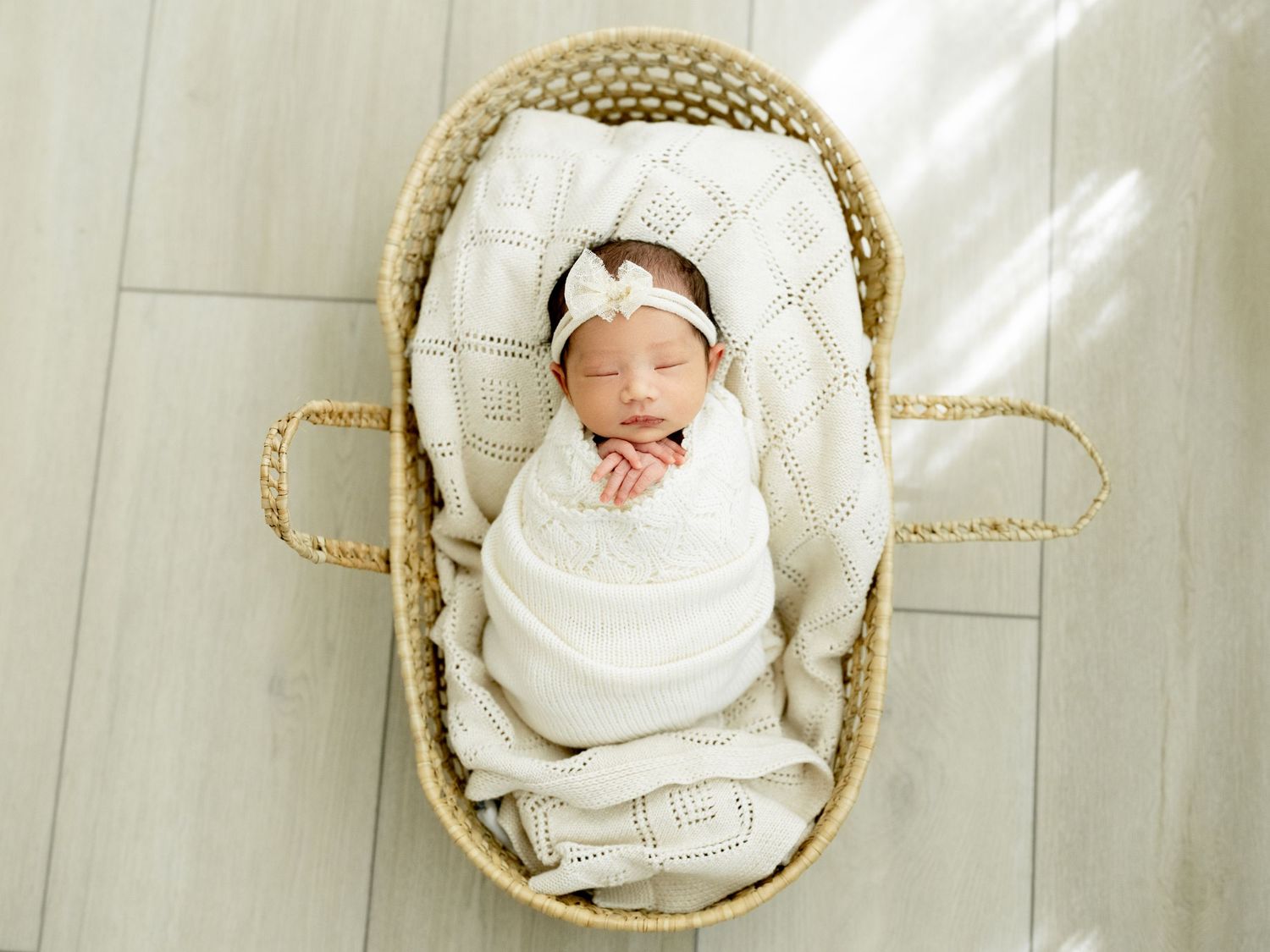 Sleeping newborn baby wrapped in white swaddle with floral headband in a wicker Moses basket.