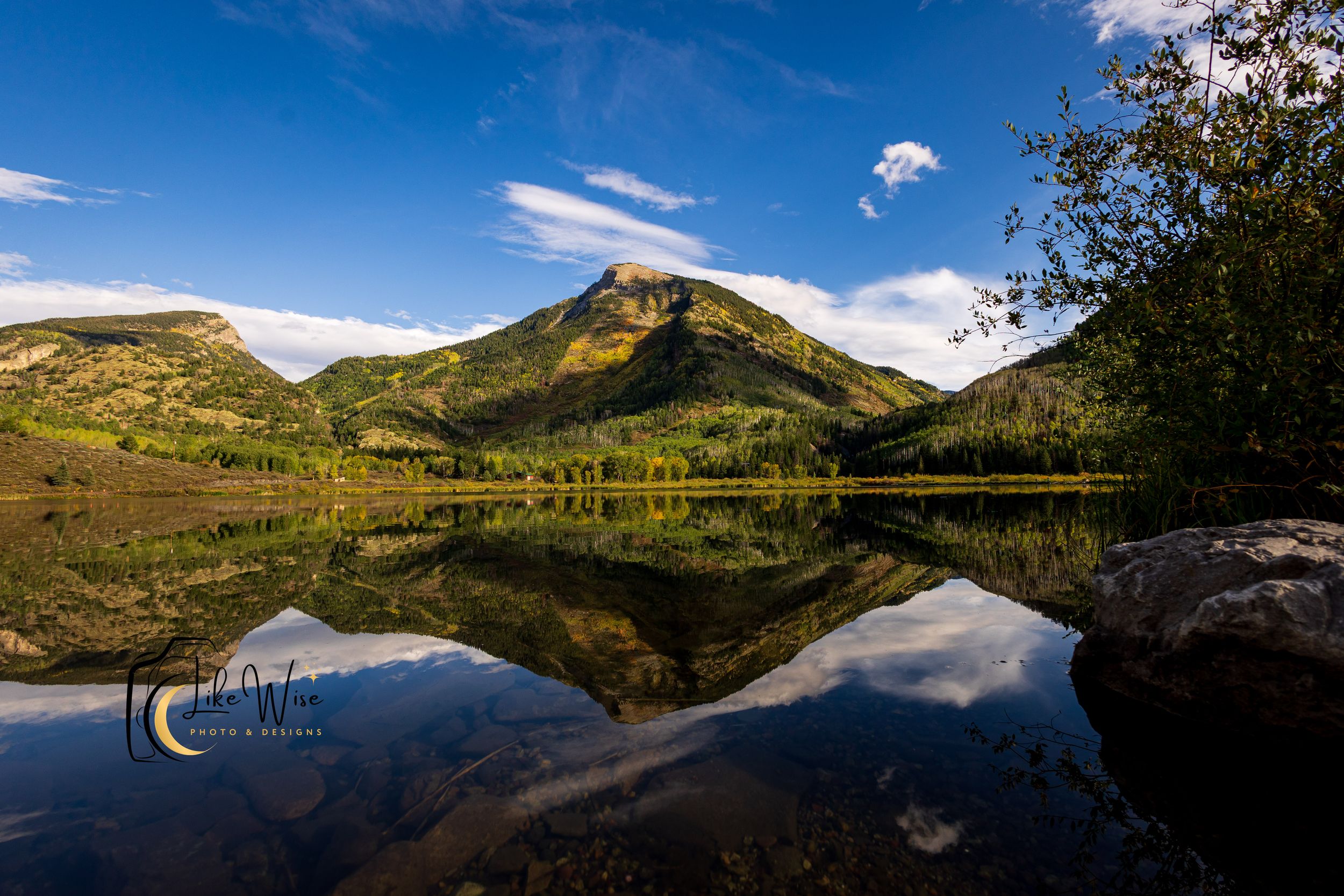 A serene mountain lake reflects a green peak and blue sky with clouds, surrounded by autumn foliage in Colorado.
