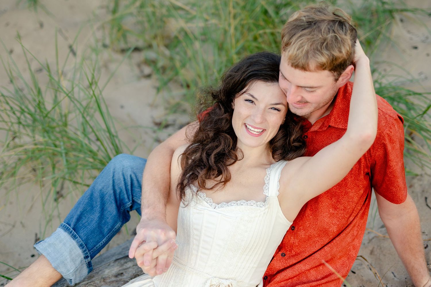 Happy couple sitting on beach sand dunes, woman in white dress smiling, man in red shirt embracing her tenderly.
