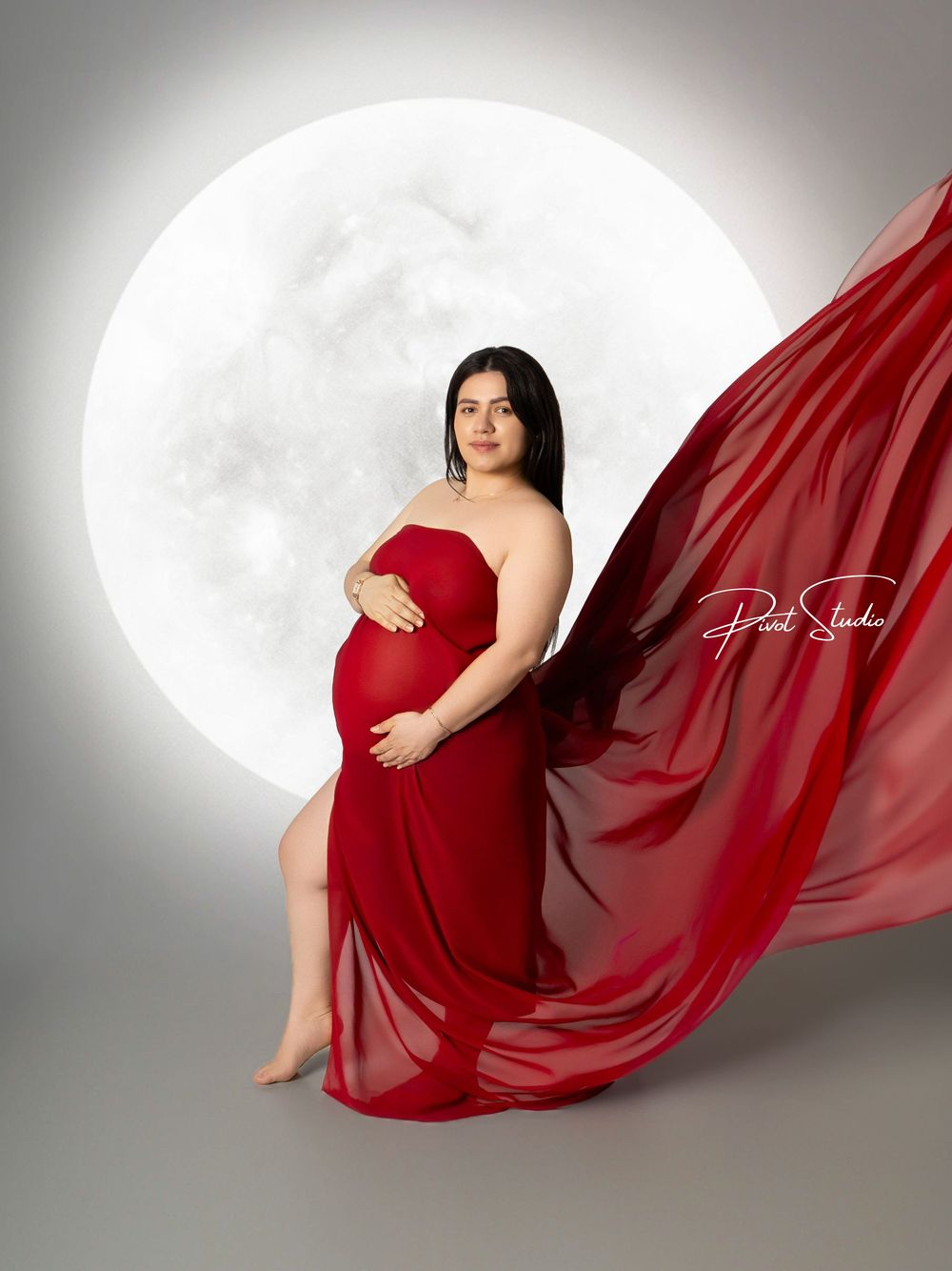 Pregnant woman in flowing red gown poses elegantly before glowing moon backdrop in professional maternity photo.