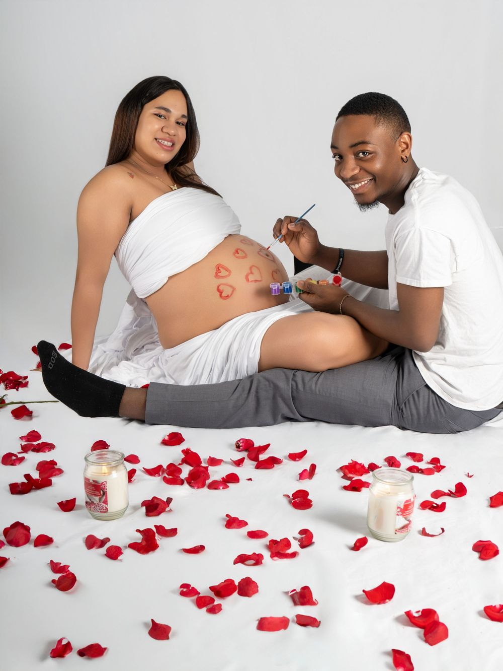 Couple doing a maternity photoshoot with rose petals, painting hearts on pregnant belly with candles nearby.