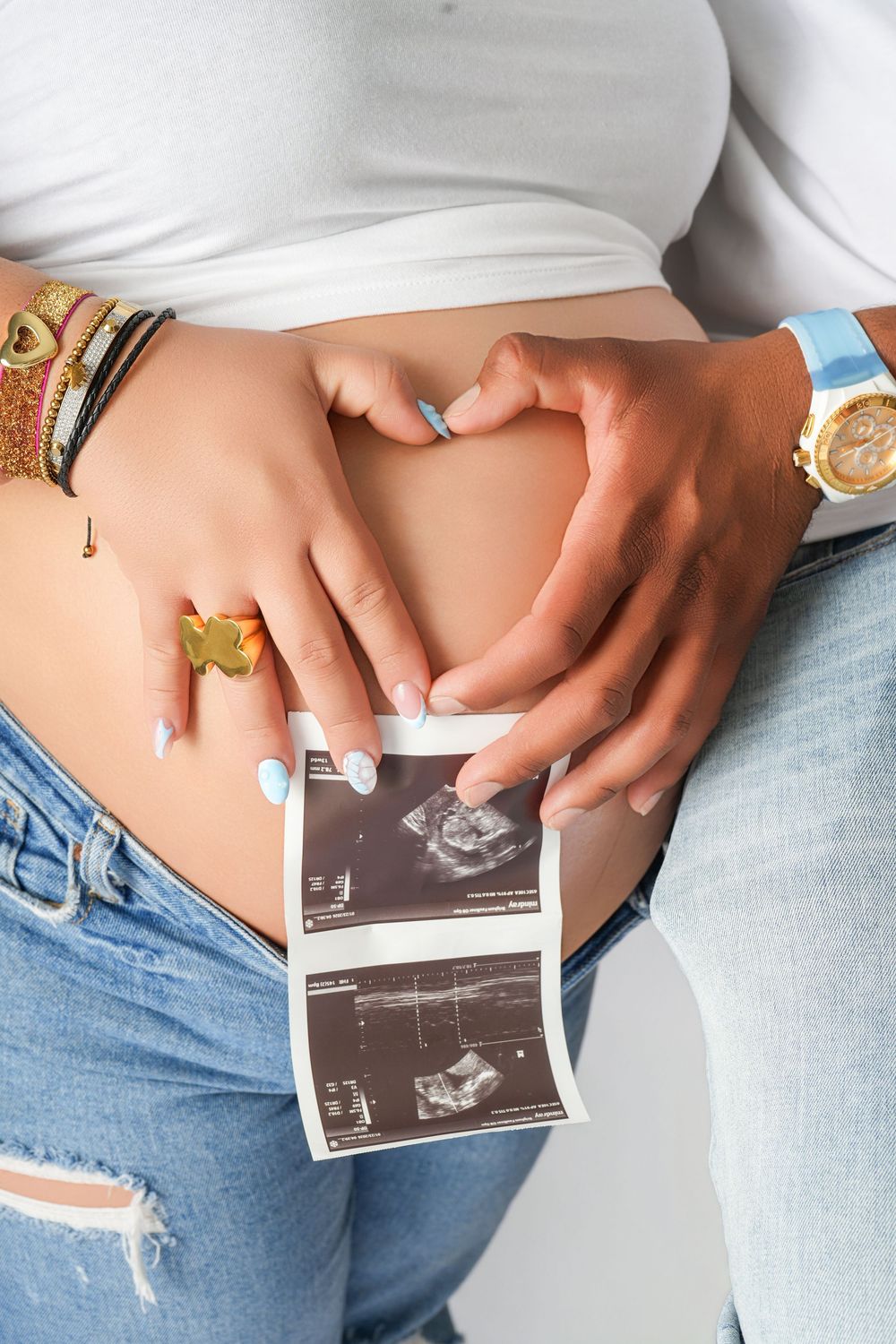 Pregnant woman holding ultrasound scan photos while forming a heart shape with hands on baby bump.
