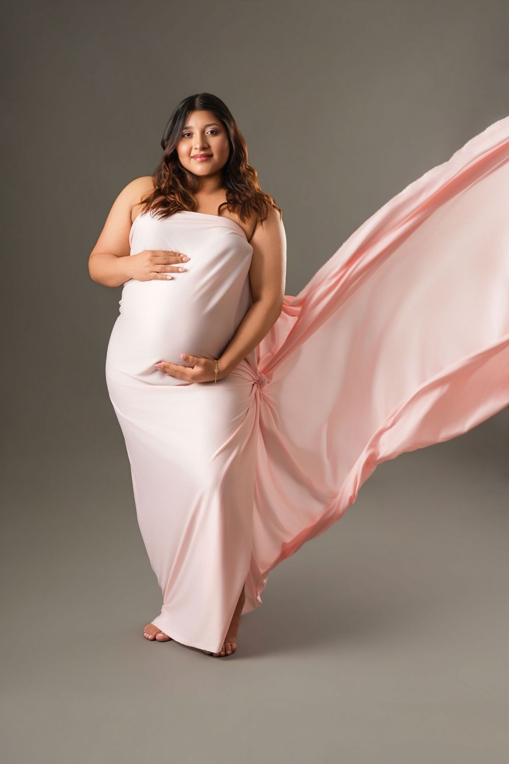 Pregnant woman in flowing pink fabric gown posing gracefully in studio maternity photography session.
