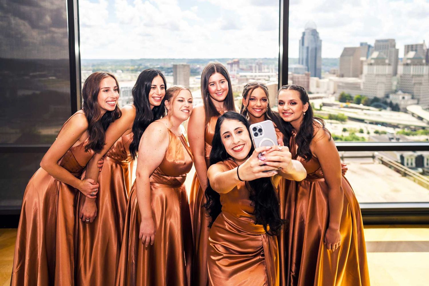 Six bridesmaids in gold satin gowns pose for a selfie near a high-rise window at the view venue