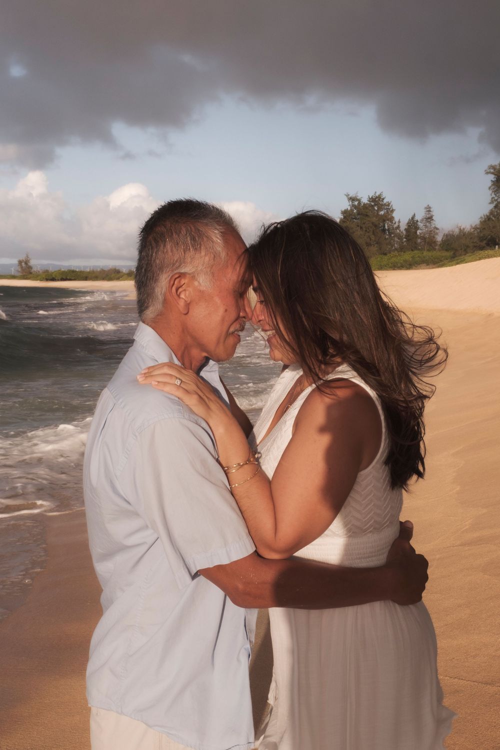 Couple embracing on a secluded North Shore Oahu beach framed by lush Hawaiian mountains during sunset.