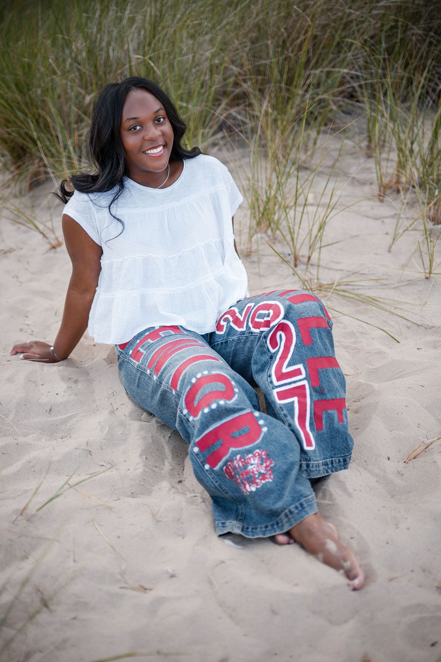 Smiling young woman sitting on sandy beach wearing white top and decorative jeans with red lettering and numbers.