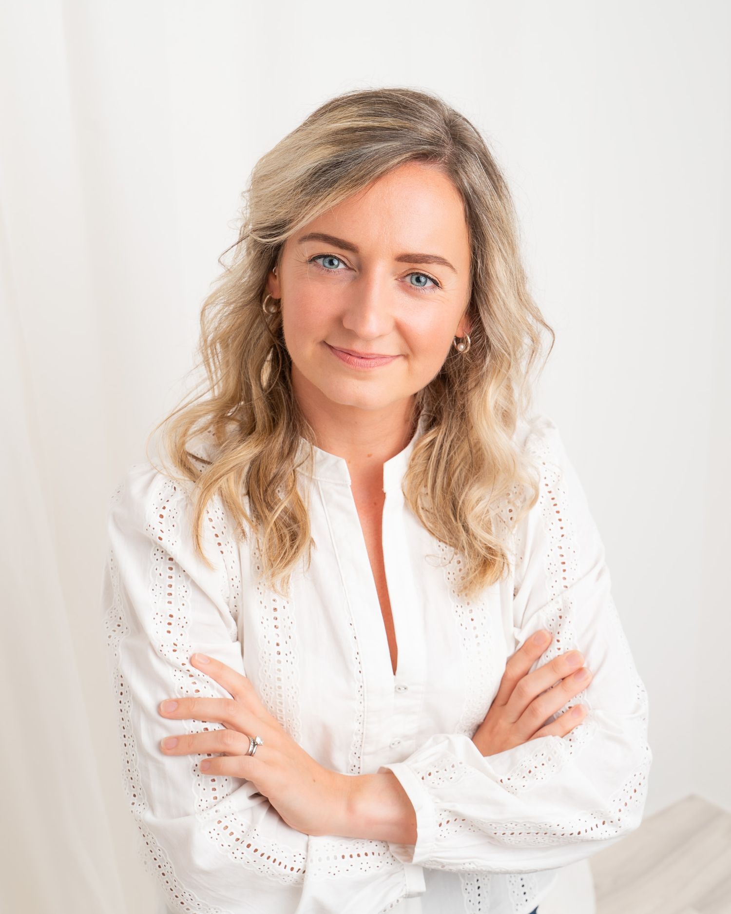 Woman with blonde wavy hair and blue eyes smiling confidently in a white blouse with arms crossed.